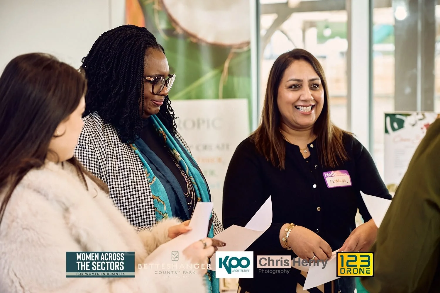 Women smiling and holding papers at an indoor networking event, with banners and logos in the background.