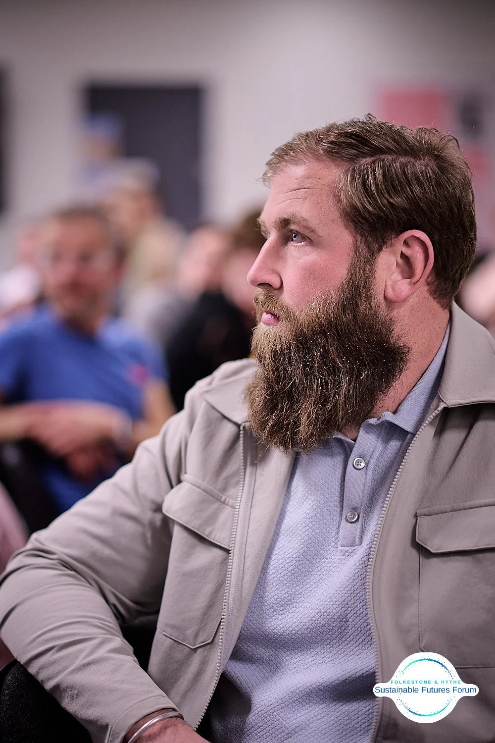 A man with a beard and mustache wearing a light gray jacket and light blue shirt, sitting at an indoor event with a focused expression, with other attendees blurred in the background.