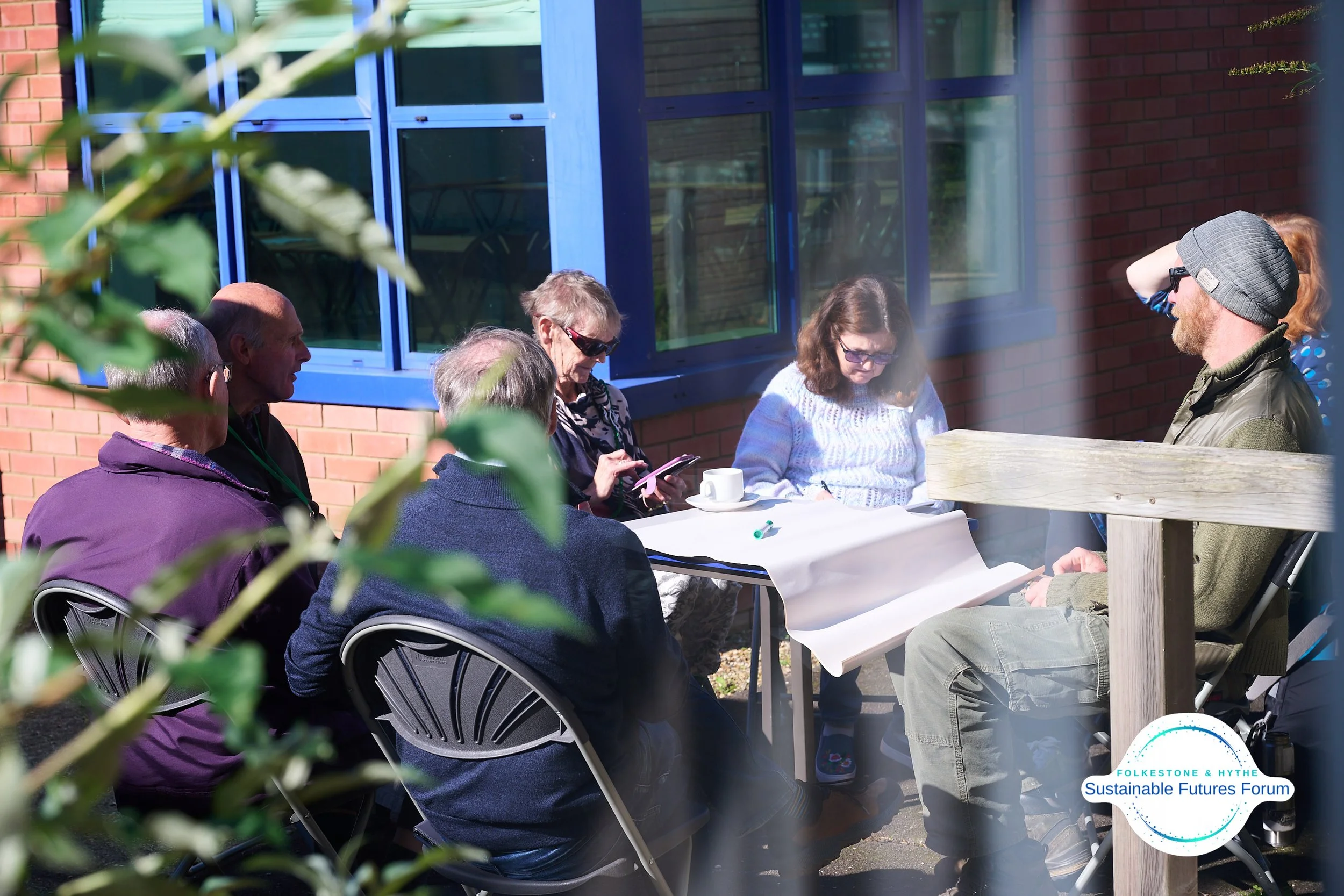 Group of six people sitting outdoors at a table, engaged in discussion, with one person taking notes and another using a phone, in front of a brick building with blue window frames, during daytime.