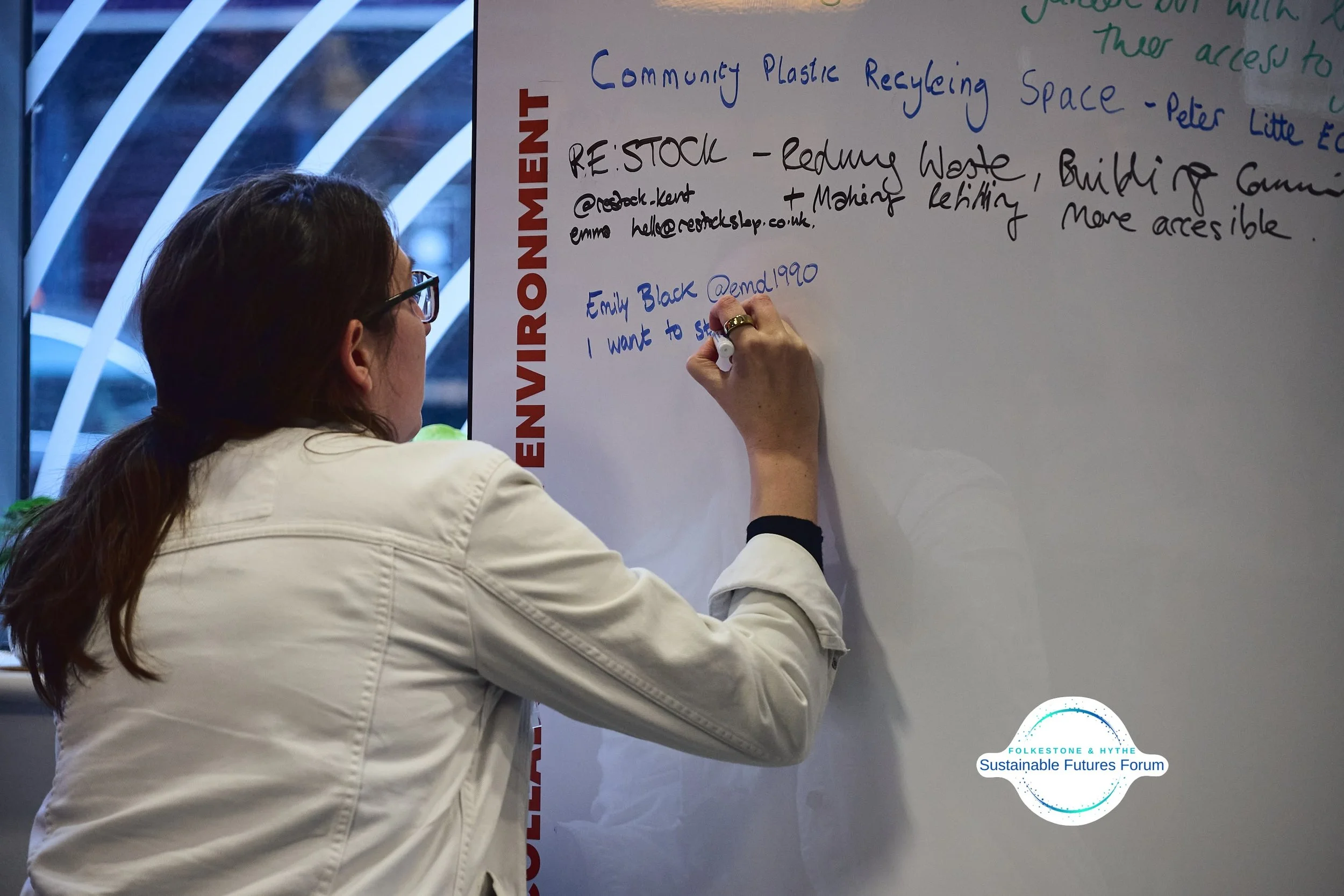 A woman writing on a whiteboard during an environmental forum, with text about community plastic recycling and reducing waste.