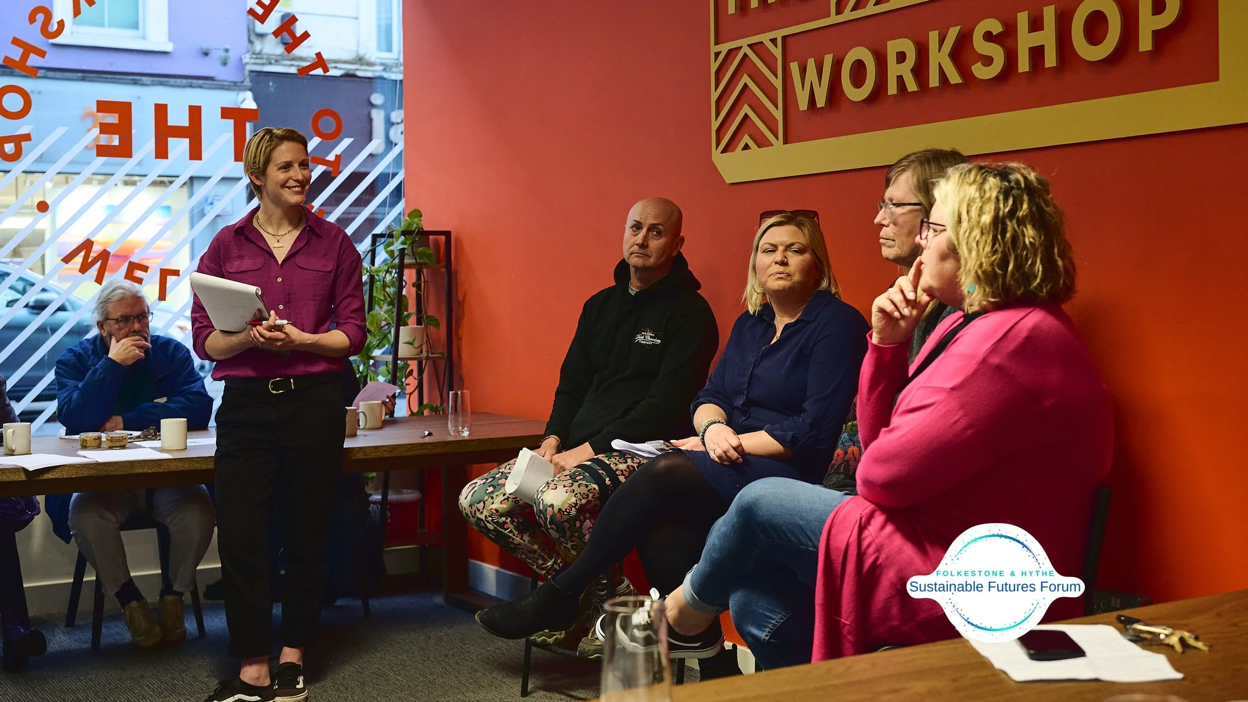 A woman standing and speaking to a seated group at a workshop, with a red wall and a sign saying 'Workshop' behind her, and a logo for Folkestone & Hythe Sustainable Futures Forum at the bottom right.