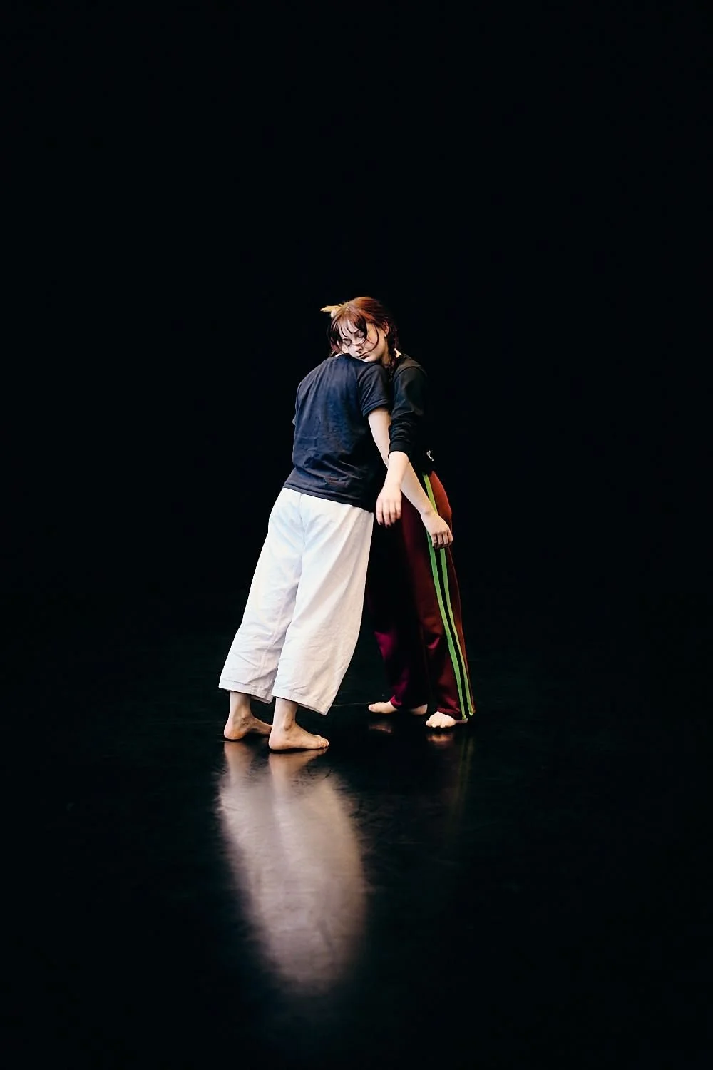 Two women practicing contemporary dance in a dark studio, with one resting her head on the other's shoulder.