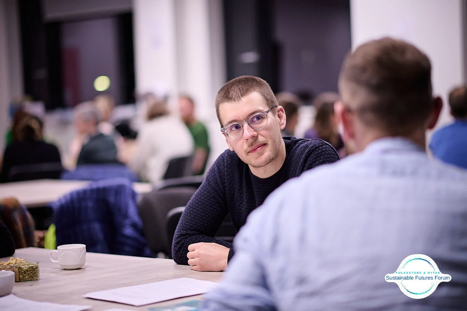 A man with glasses and a black sweater listening to another person in a conference room filled with attendees, tables, and papers.