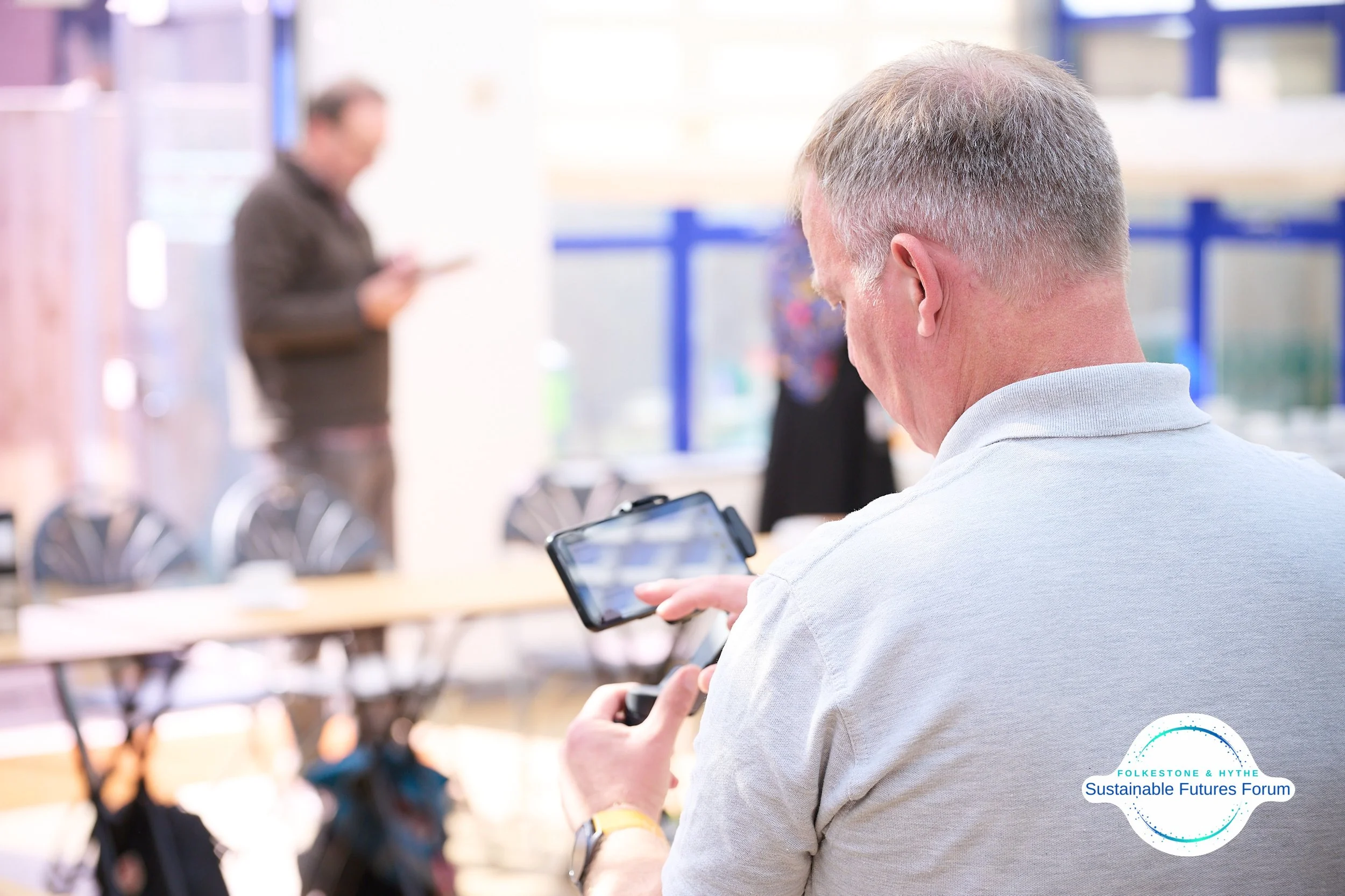 Man looking at smartphone in a conference room with blurred individuals in the background, and a logo that says 'Folkescone & Hythe Sustainable Futures Forum' in the bottom right corner.