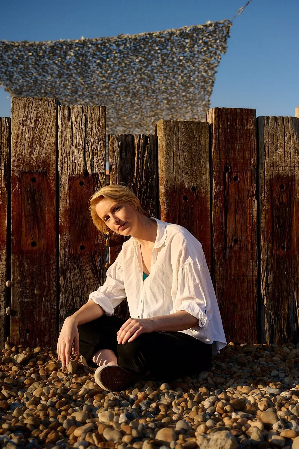 A woman with blonde hair sitting on a rocky beach in front of a wooden fence, with a netted shade above, during sunset.