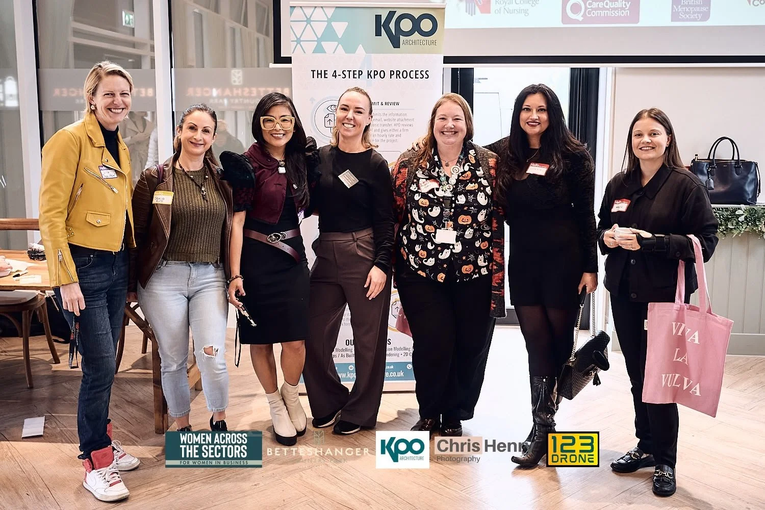 Group of seven women standing together indoors at a professional event, smiling at the camera. Behind them are promotional banners and a large screen displaying logos and information.