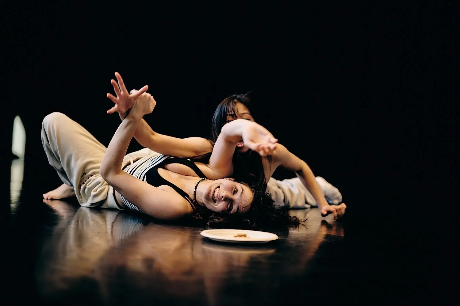 Two women are performing a dance or theatrical act on a dark stage, one lying on the floor and smiling, the other leaning over and appearing to interact.