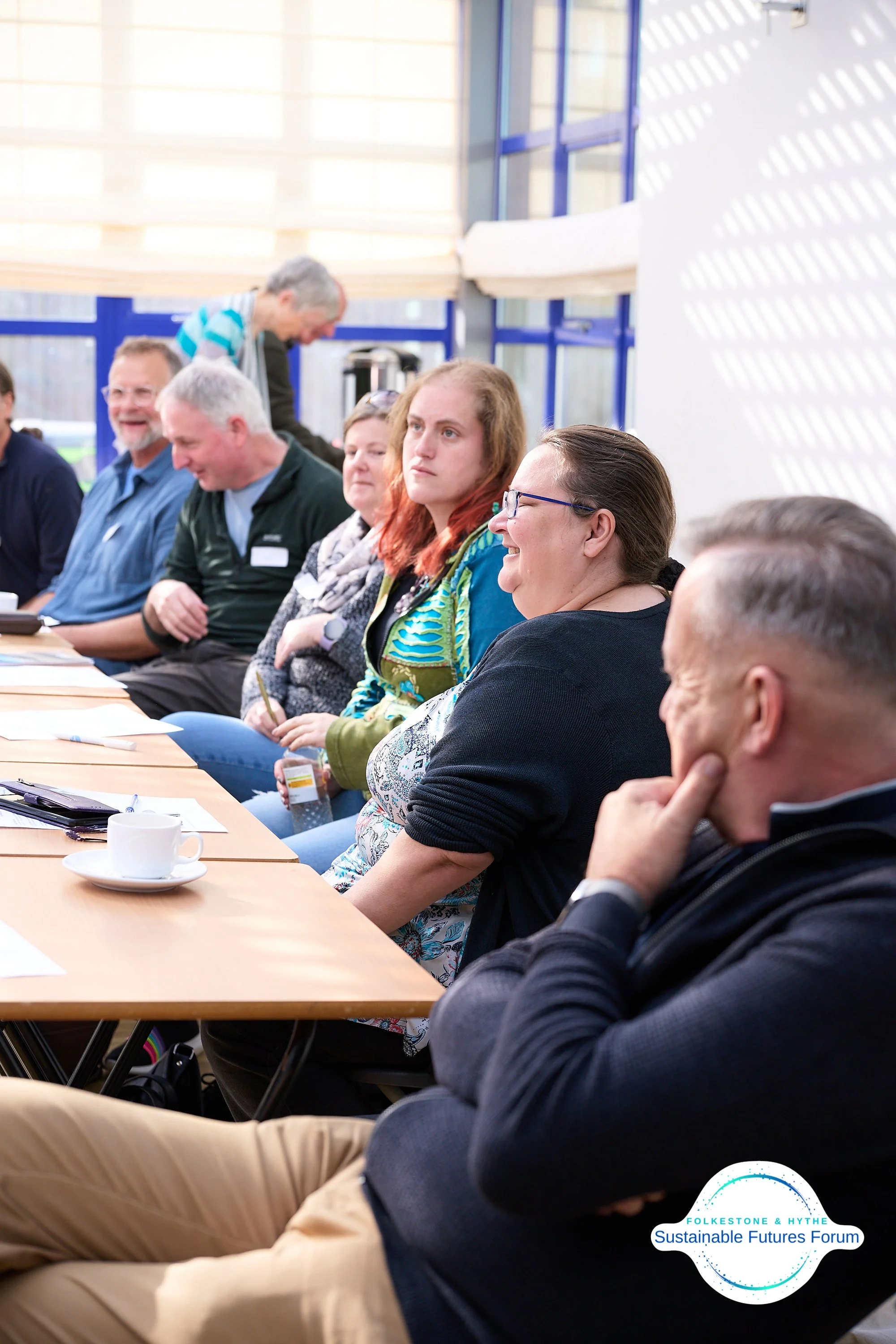 People seated at a long table during a forum or workshop, with some taking notes and a logo on the bottom right that reads 'Folkestone & Hythe Sustainable Futures Forum'.