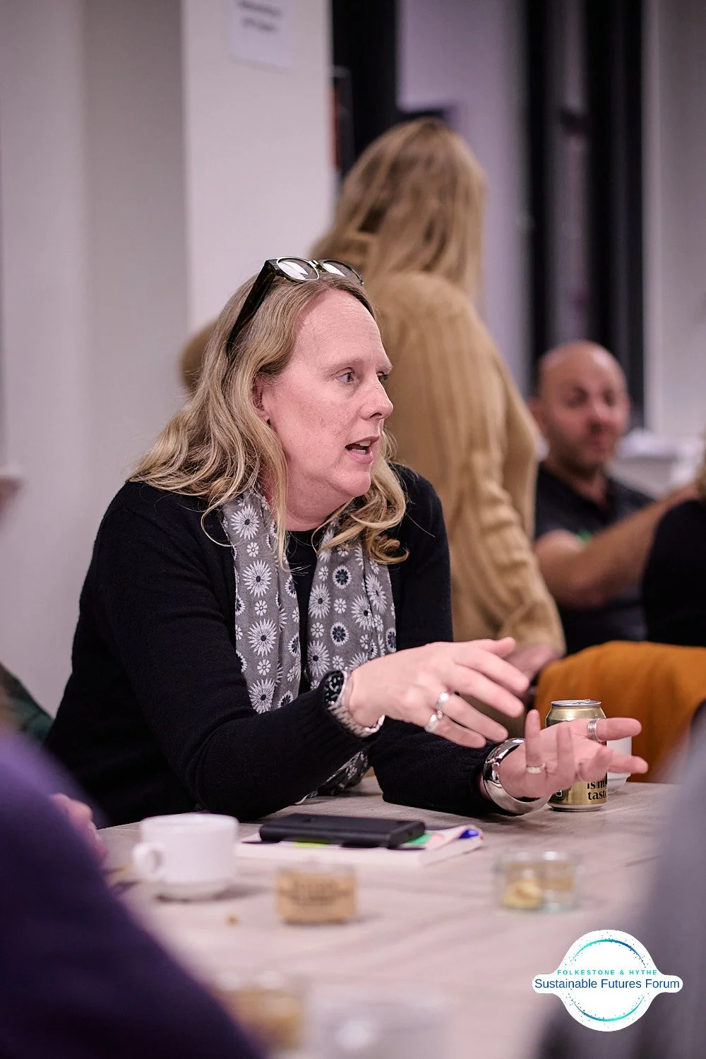 A woman with blonde hair and glasses on her head, wearing a black top and a gray scarf with white flowers, is speaking and gesturing during a discussion at a meeting. On the table in front of her are a smartphone, a cup, and some snacks. In the backg