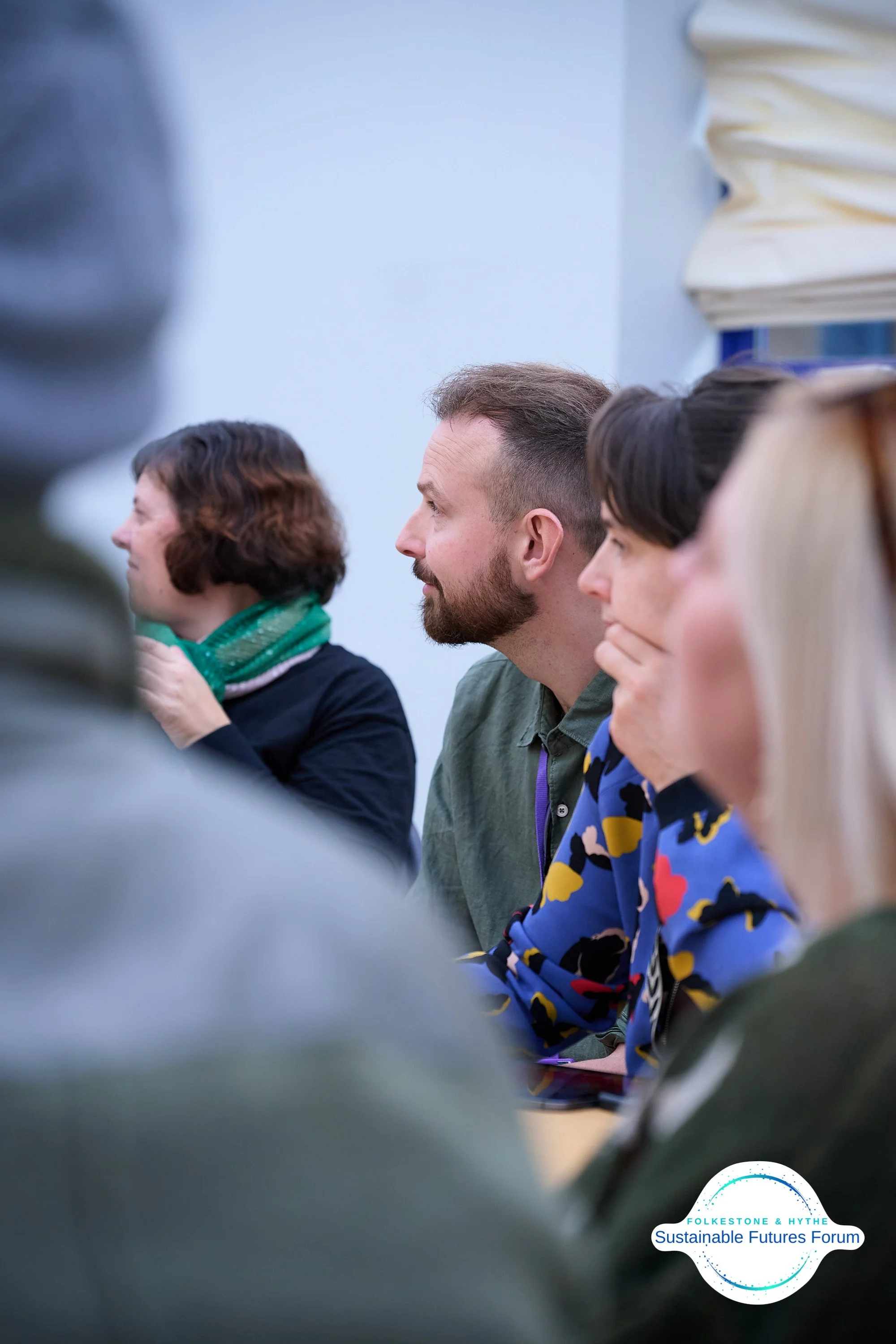 People attending a forum or conference, sitting in a row, listening attentively. A logo in the bottom right corner reads 'FOLKESTONE & HYTHE Sustainable Futures Forum.'