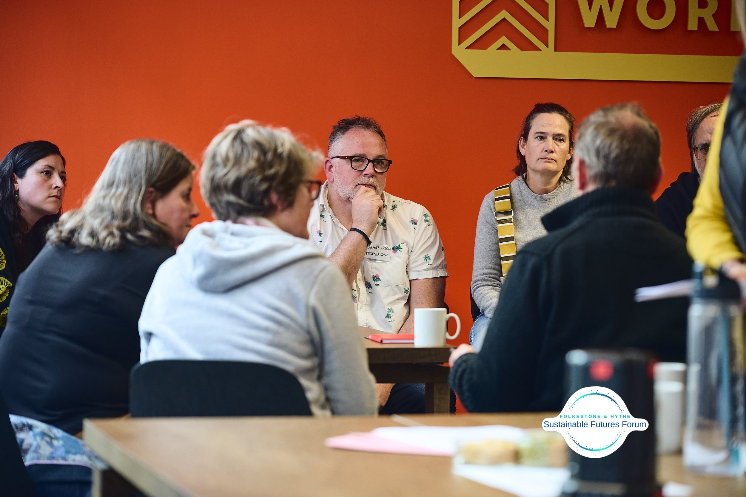 Group of people attending a meeting at the Sustainable Futures Forum in Felkstene & Hythe, sitting around a table against an orange wall.