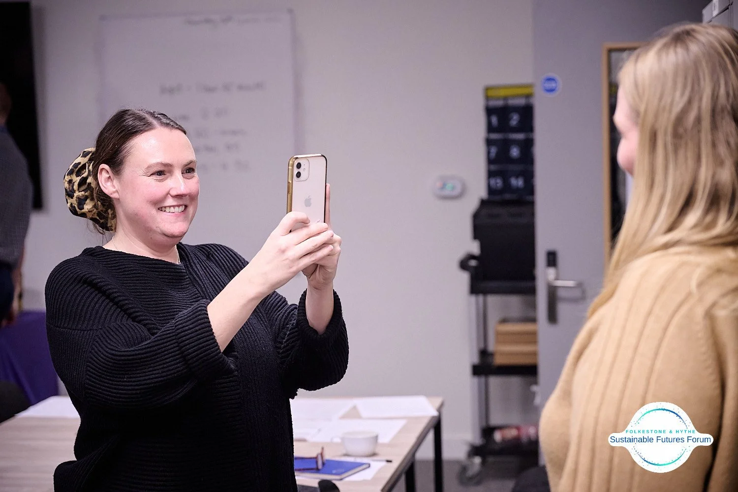 A woman with a leopard print hair accessory taking a selfie with another woman in an indoor setting.