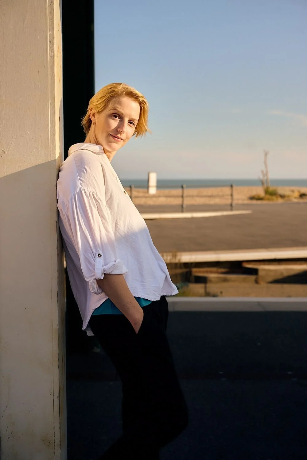A woman with blonde hair in a white shirt and black pants leaning against a wall near a beachside road at sunset.