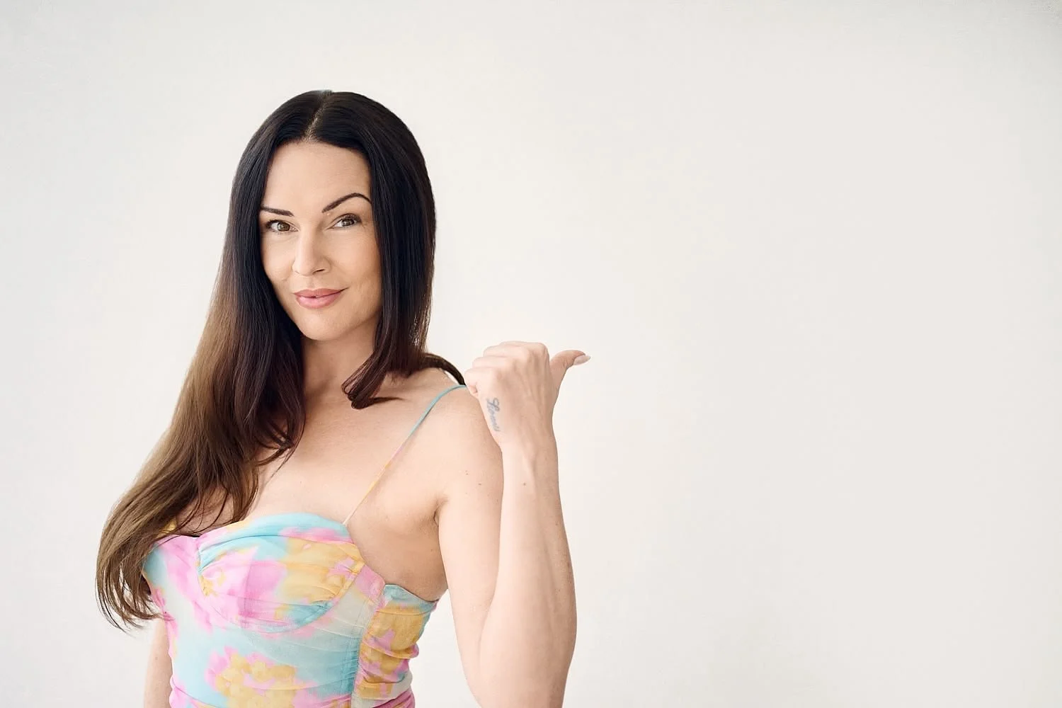 A woman with long dark hair wearing a colorful floral dress, smiling and pointing at her shoulder with her thumb.