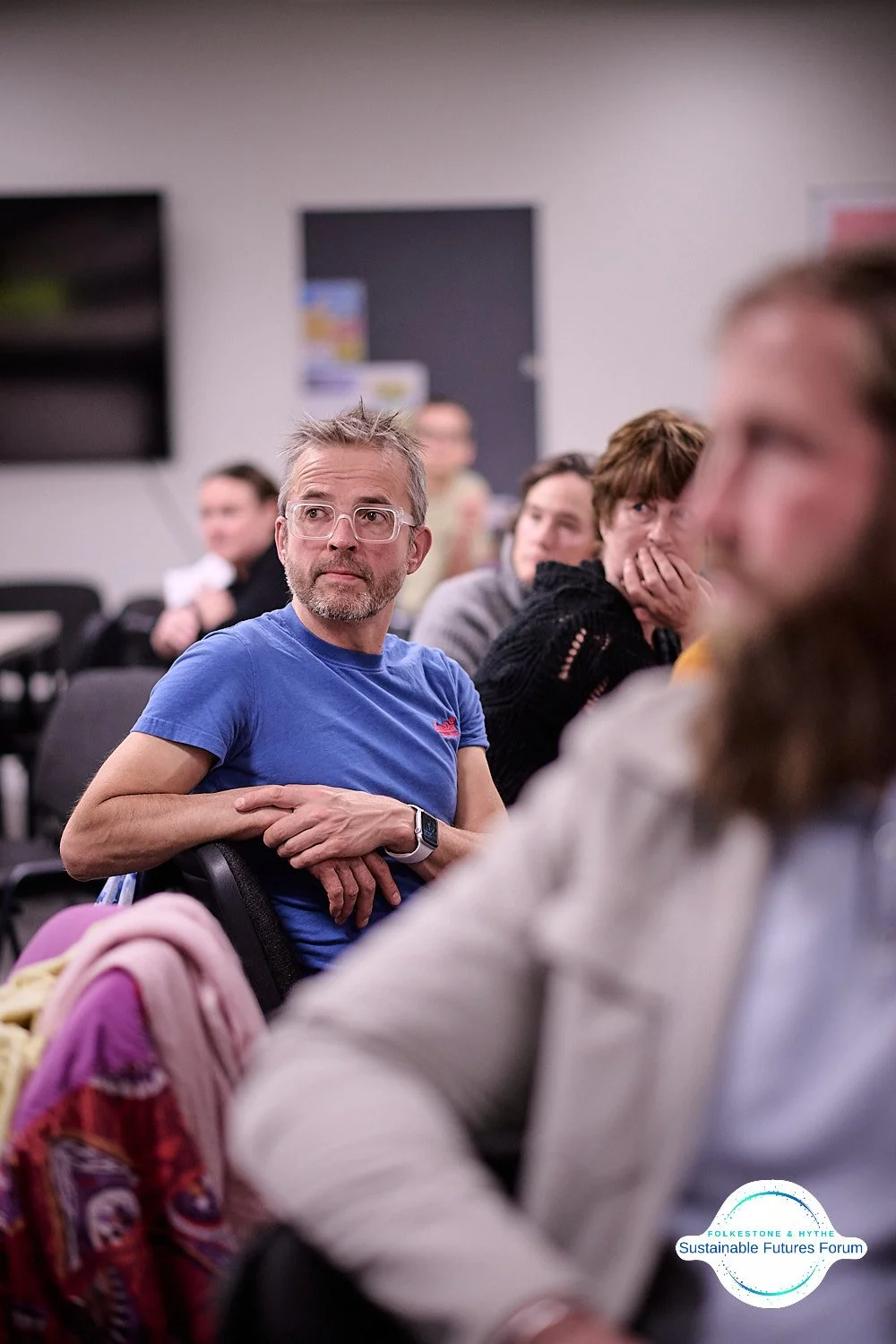 A group of people attending a forum, with a man in a blue t-shirt and glasses looking attentively, seated in a room with a white wall and a black television screen in the background.