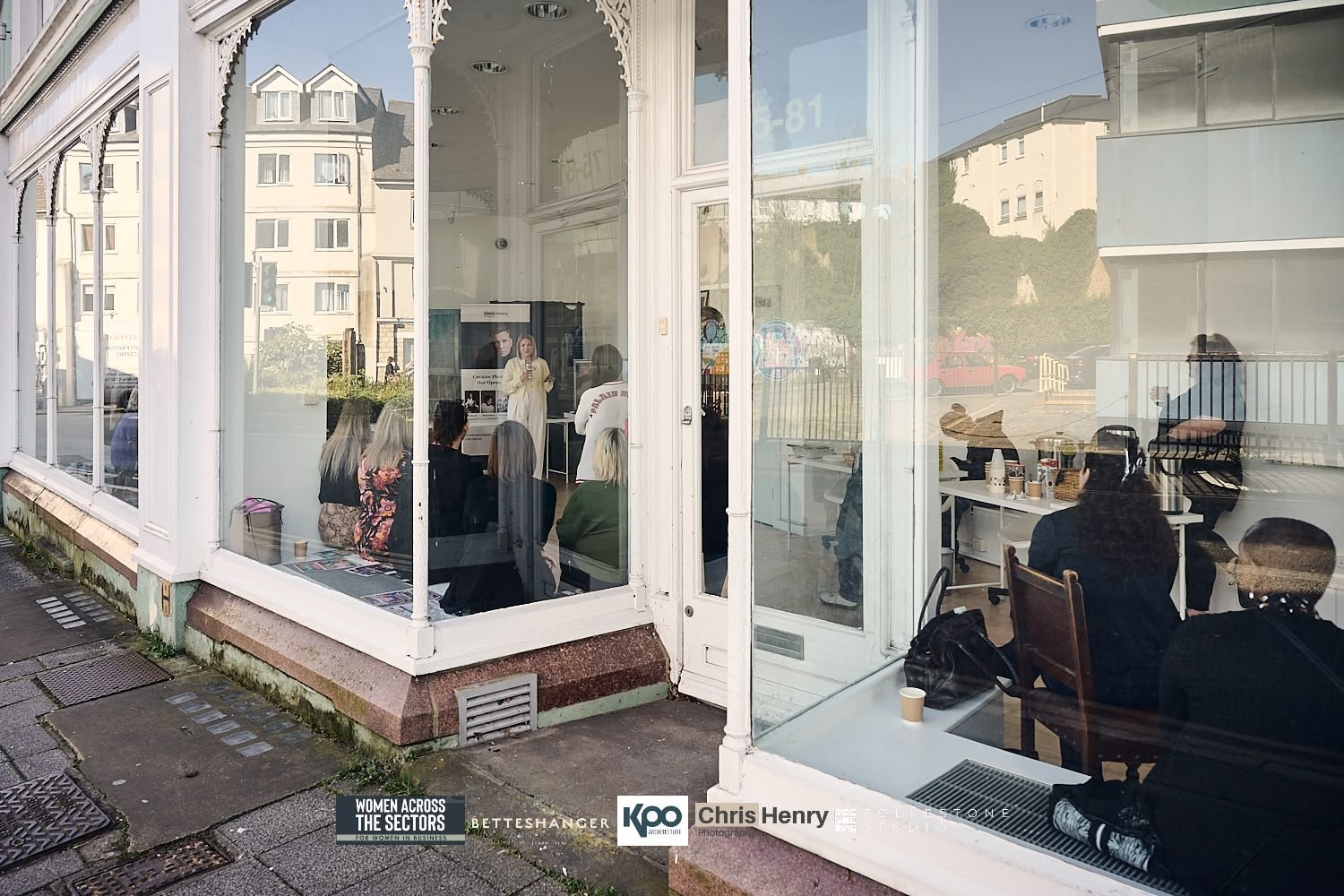 A group of women attending a presentation inside a white-framed glass storefront, with some seated and one woman speaking at the front. Reflections of outdoor buildings and parked cars are visible on the glass.