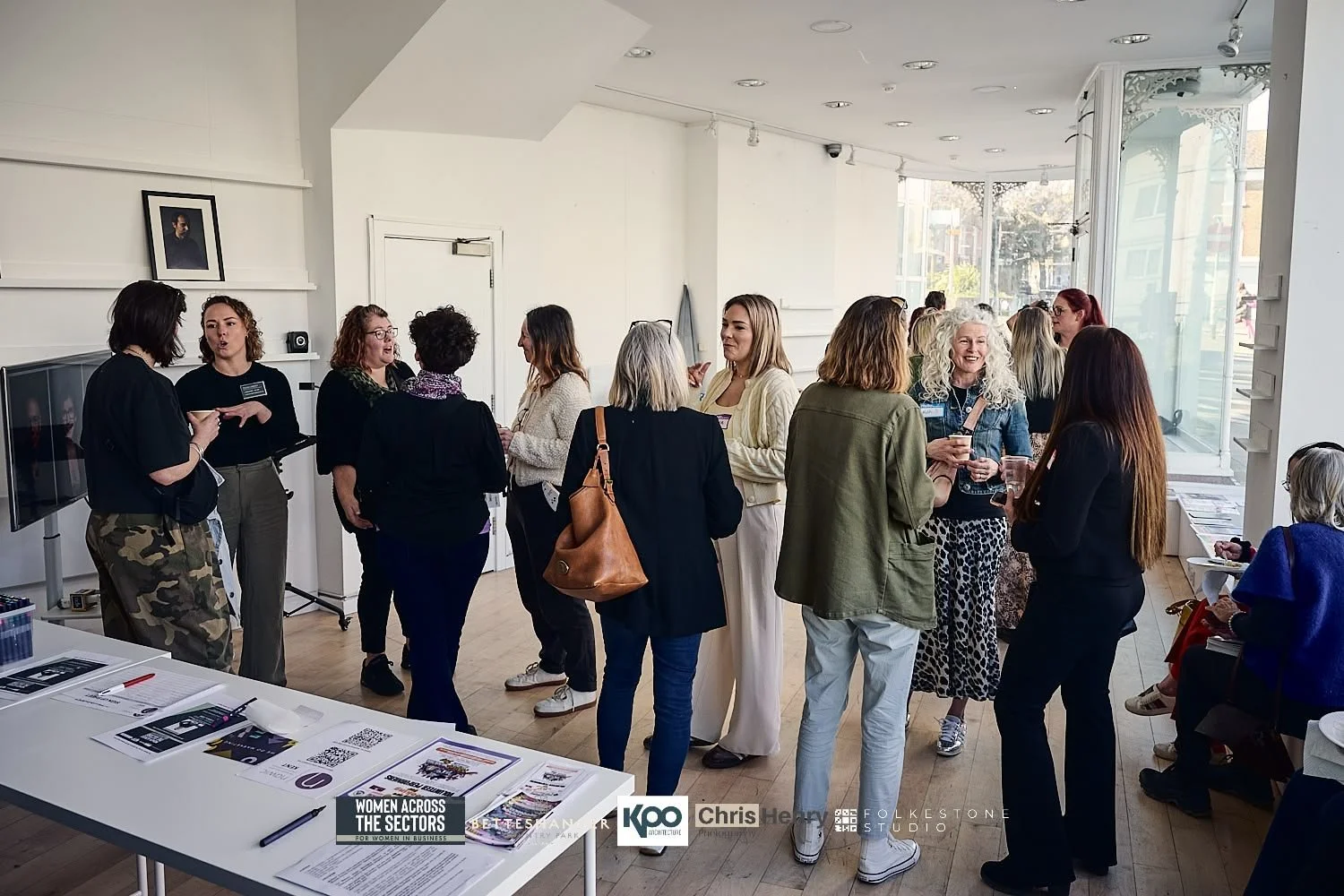Women networking and chatting at an indoor event or conference, with informational flyers on a table in the foreground.