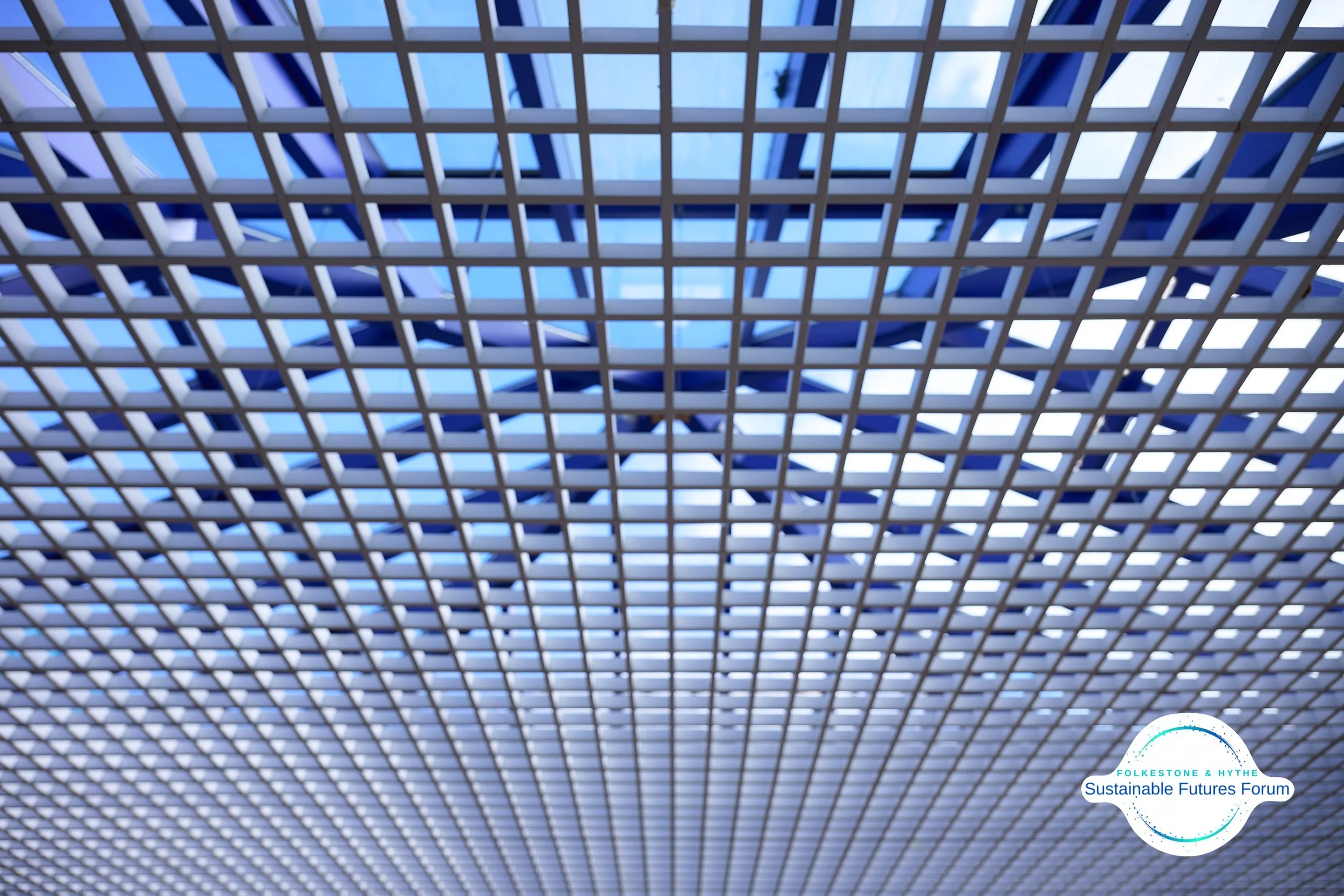 Close-up view of a modern architectural ceiling with a grid pattern and blue accents, featuring a logo for Folkesone & Hythe Sustainable Futures Forum in the bottom right corner.