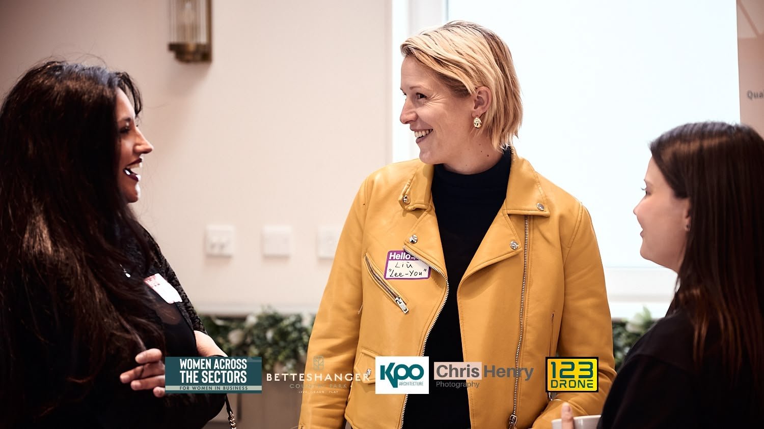 Three women engaged in conversation at a professional event, with one woman wearing a yellow jacket and all smiling.