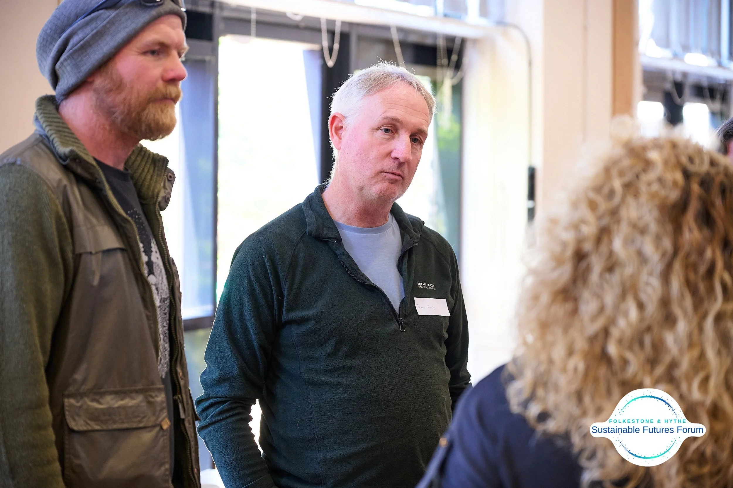 A group of people attending a forum or discussion, with two men visible in the foreground, one with a beanie and the other with short grey hair, engaging in conversation with a woman with curly blonde hair. A logo for FolkesTone & Hythe Sustainable F