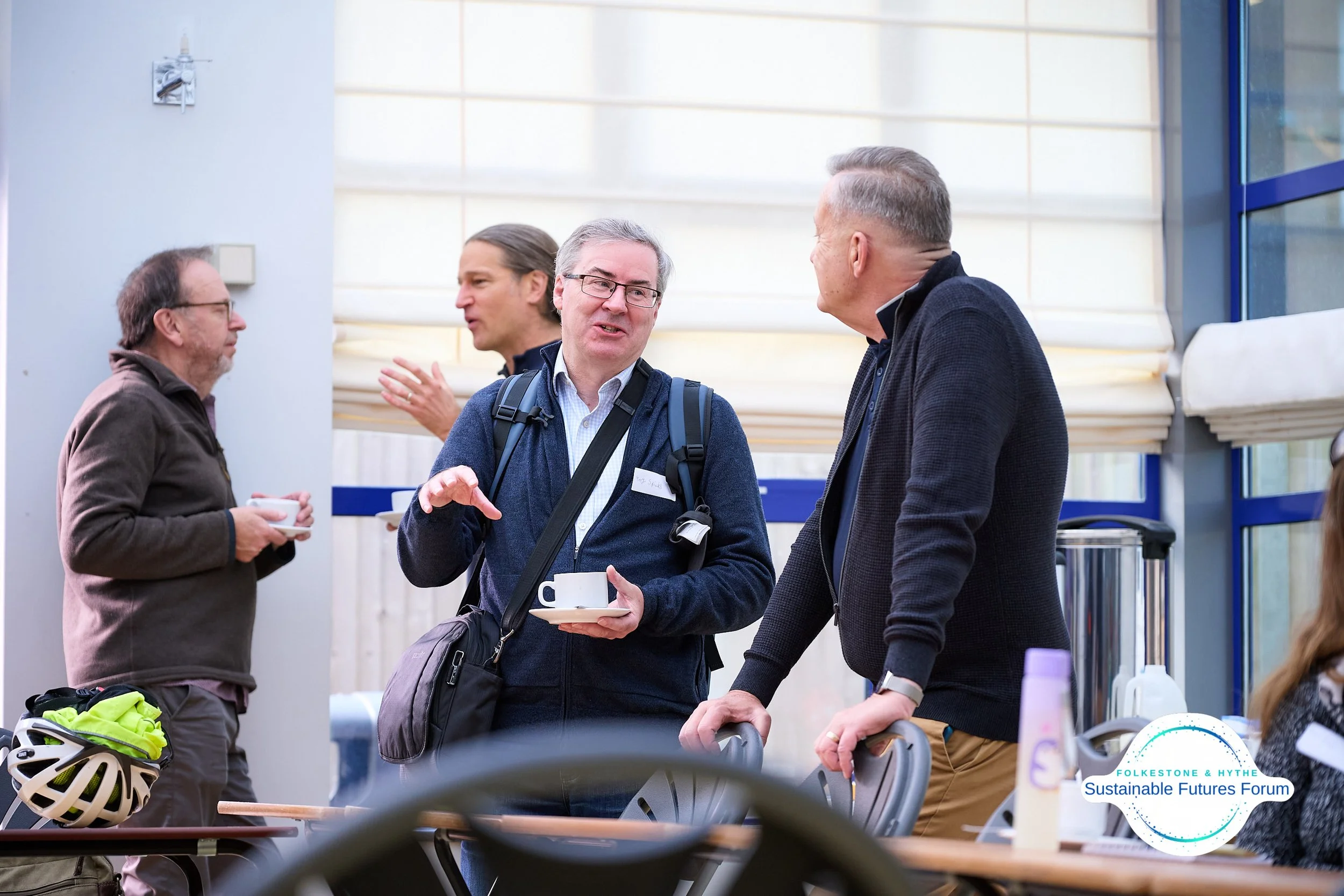 Four men are engaged in conversation in a conference or workshop setting. Three are standing, one is partially visible, all holding cups, in a room with large windows and a sign that reads 'Sustainable Futures Forum'.