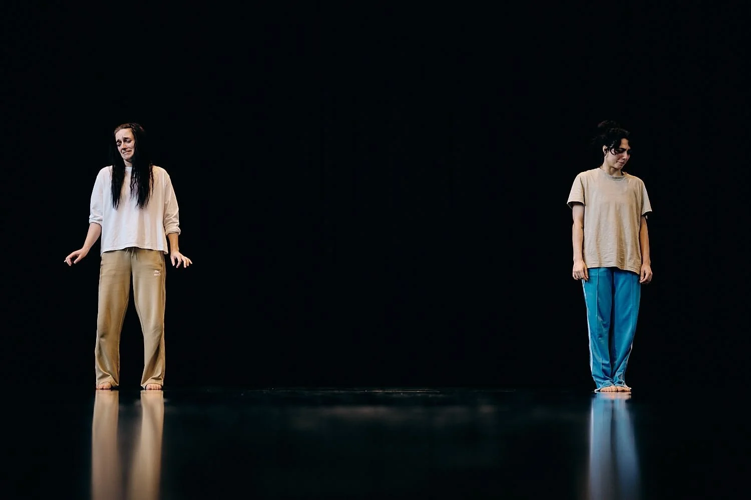 Two women stand apart on a dark stage, wearing casual clothing and barefoot, one looking distressed and the other looking downcast.