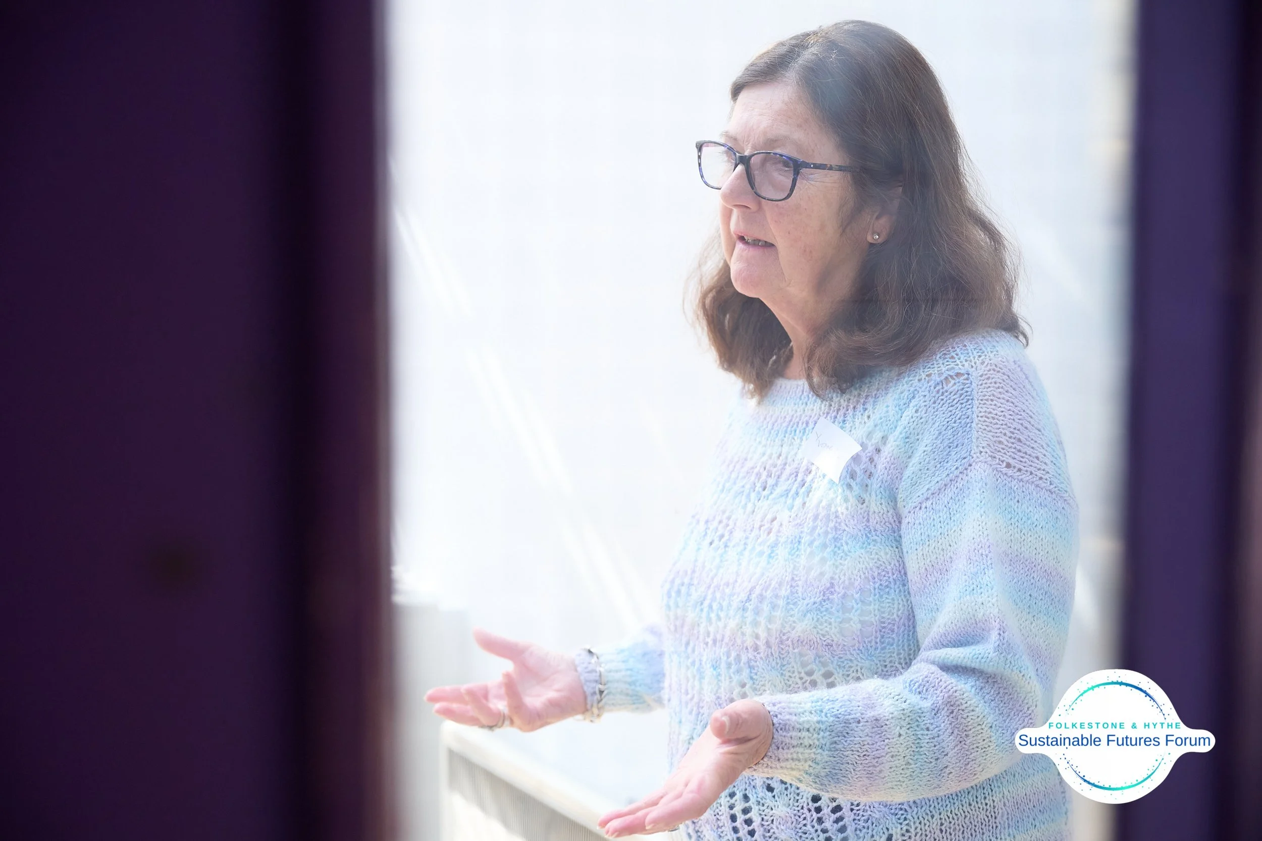 Woman with shoulder-length hair and glasses speaking, wearing a pastel-colored sweater, participating in a presentation or discussion, with the logo of Folkesone & Hythe Sustainable Futures Forum in the bottom right corner.