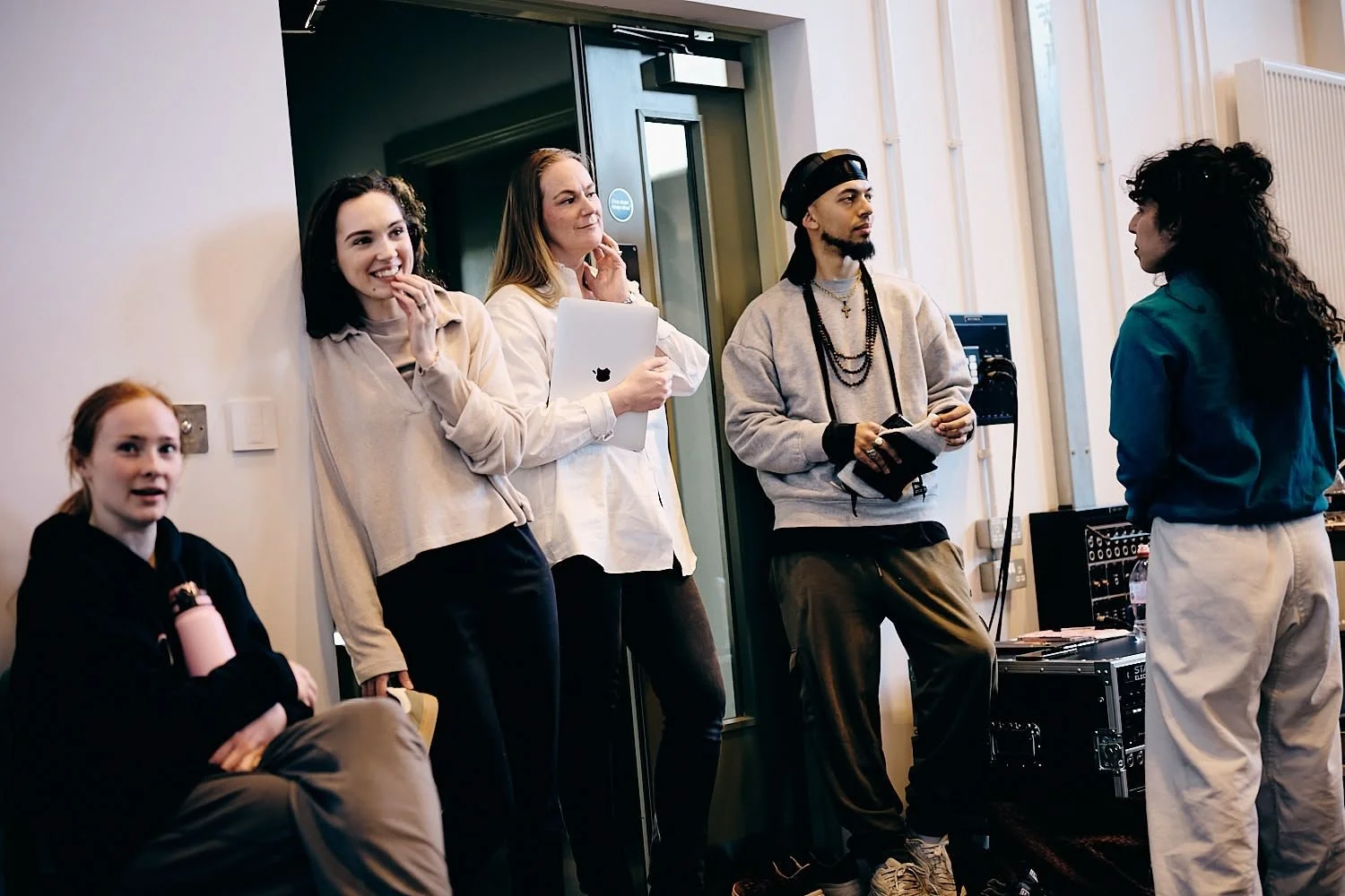A group of five people in a room having a conversation. Two women are standing, and one woman and two men are sitting or leaning against the wall. One woman holds a folder, and one man has headphones and a notebook. The setting appears to be a casual