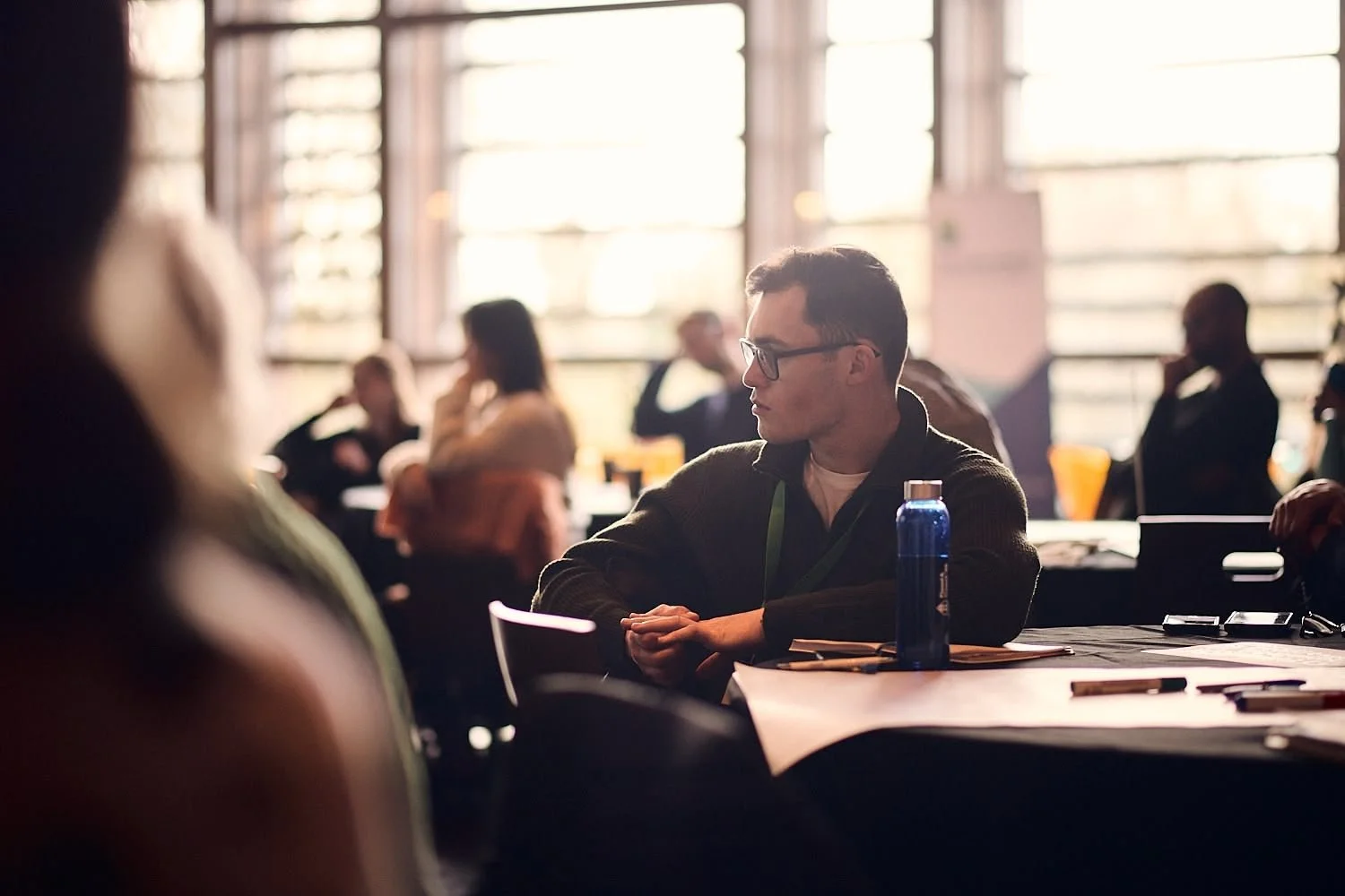 A young man with glasses and a dark hoodie seated at a table in a brightly lit room with large windows, attending a gathering or conference, with others seated around.