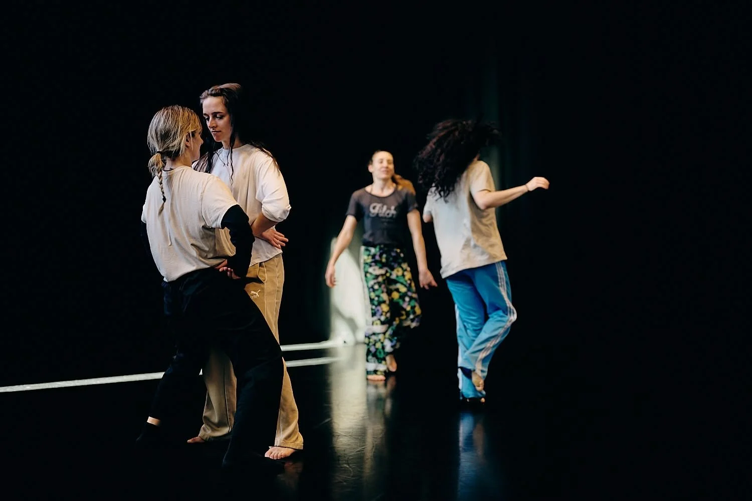Group of women practicing dance or theater on stage with black background, some engaged in conversation, others dancing or walking.