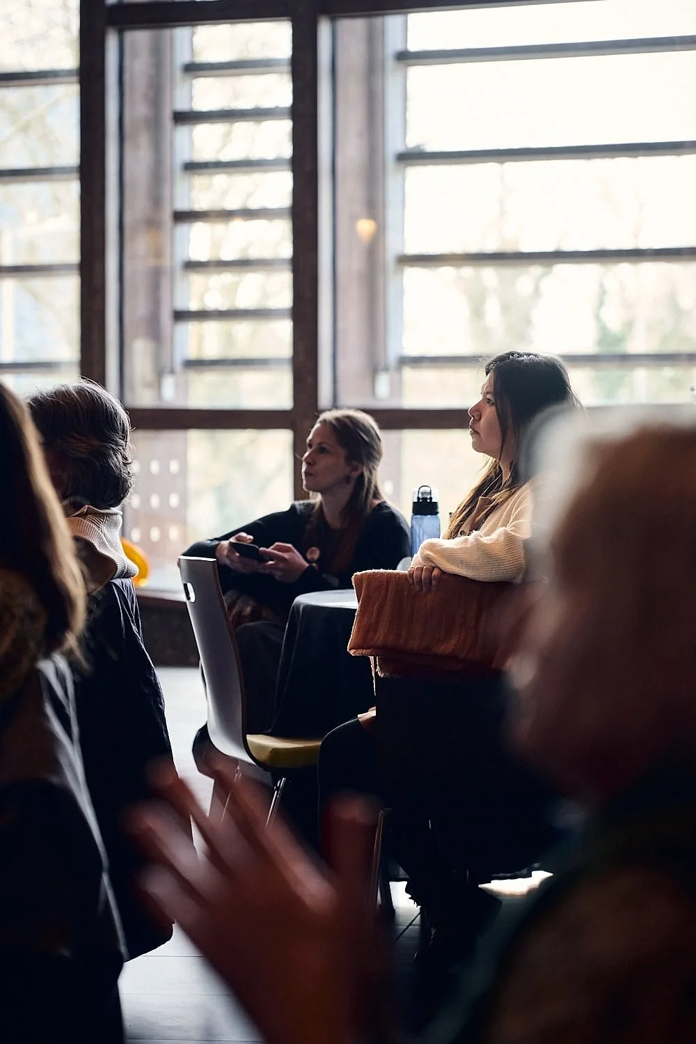 Two women sitting at a table in a room with large windows, engaged in a conversation, with other people visible in the foreground.
