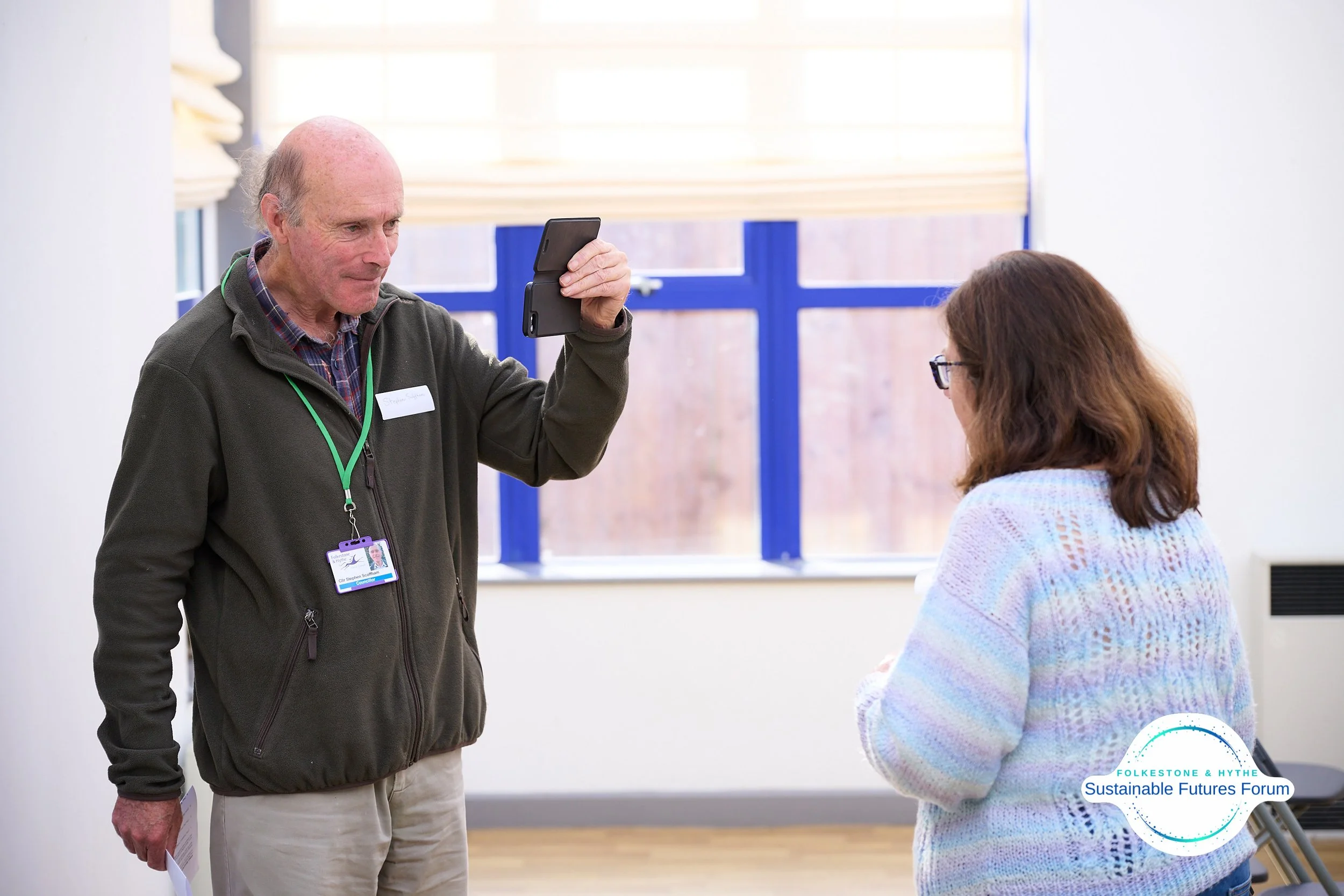 A man with a badge and lanyard taking a photo of a woman with glasses, in a room with large windows and a logo for the Folkesone & Hythe Sustainable Futures Forum.