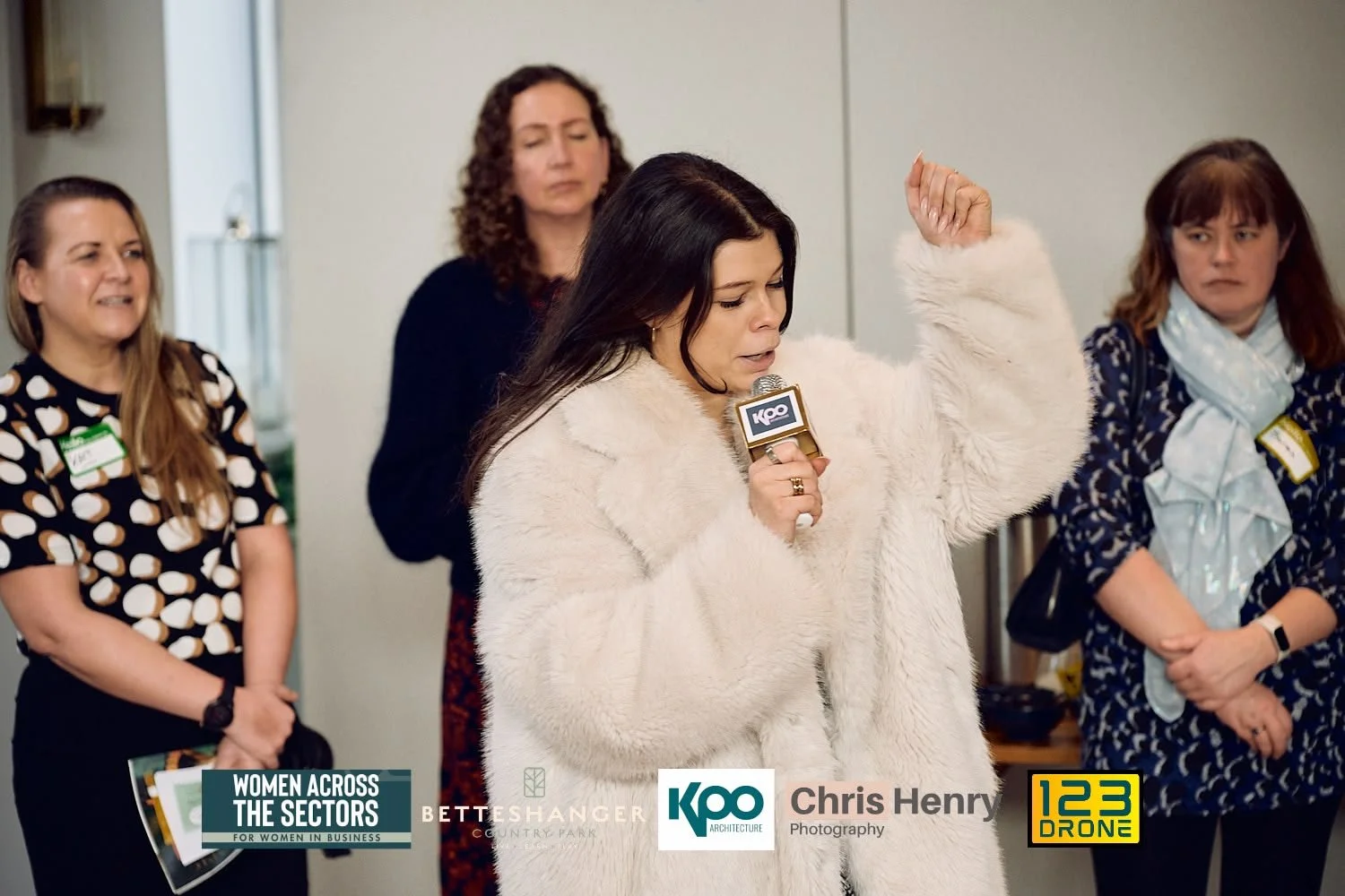 A woman in a white coat speaking into a microphone at a women's business event, with three women listening in the background.