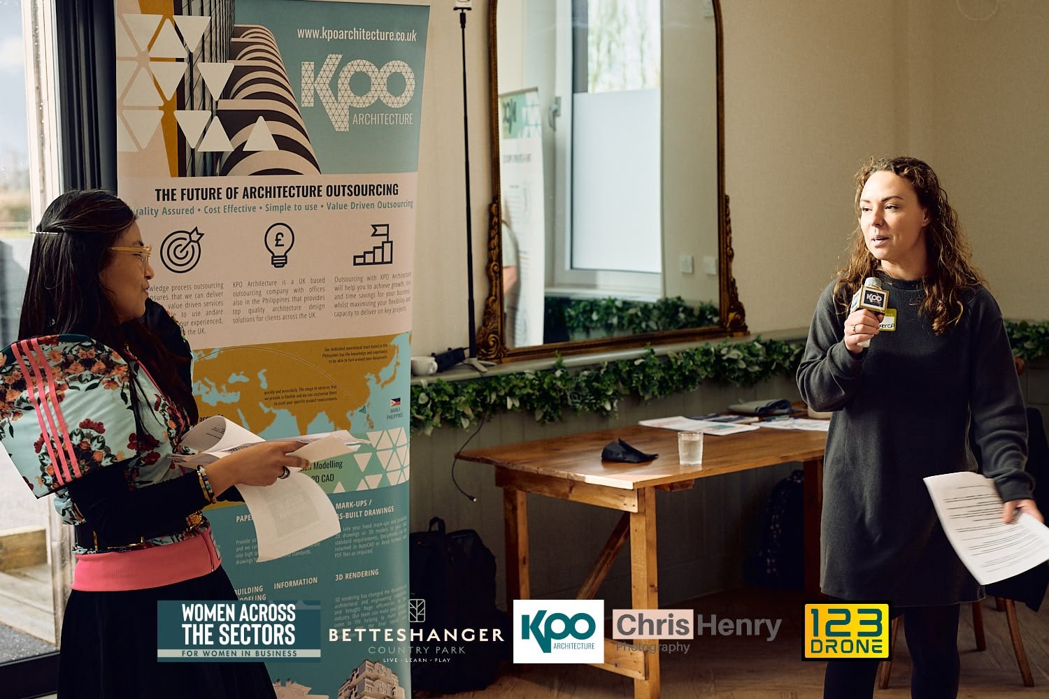 Two women speaking at a business event with a large informational poster about architecture outsourcing, in a room with a window and a wooden table.