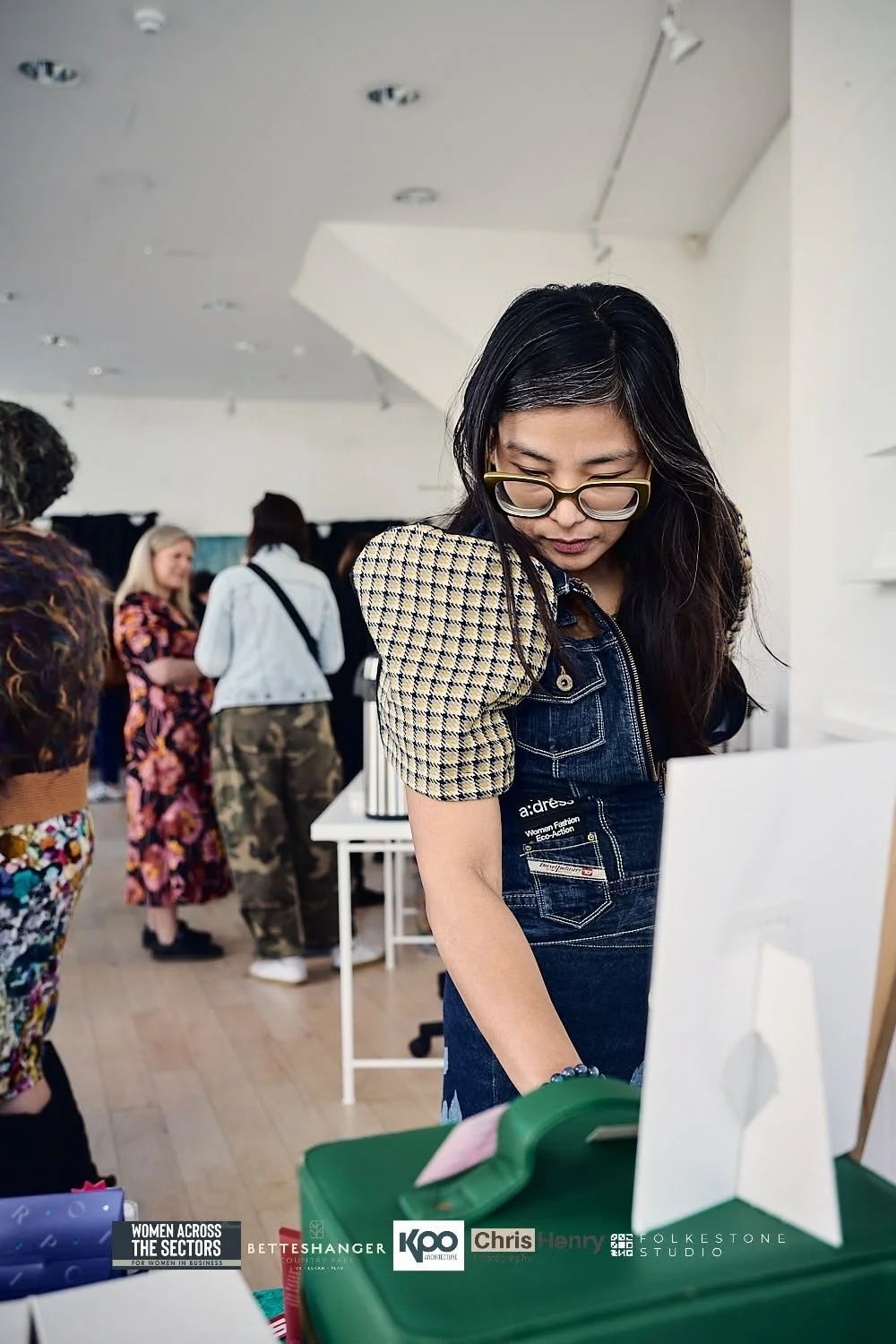 A woman with glasses looking down at merchandise in a store or event space with other women in the background.
