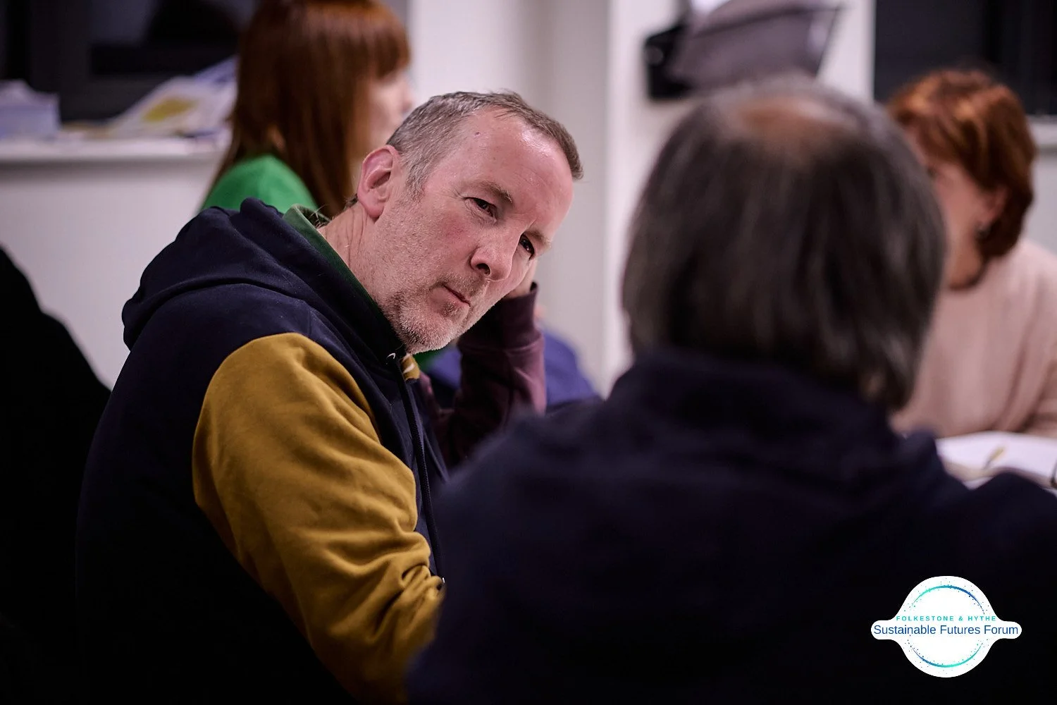 A man with gray hair and a beard in a black and yellow hoodie, sitting at a table during a meeting or discussion, looking intently at someone off-camera. In the background, other people are engaged in conversation or taking notes. There is a logo at 