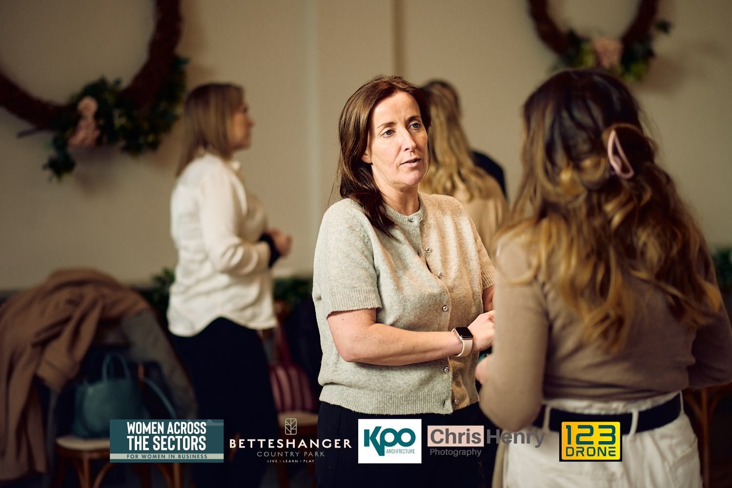 Women having a conversation at a networking event, with many women in the background, and logos for Women Across The Sectors, BETT SHANGER, KAO Architecture, Chris Henry Photography, and 123 Dron in the bottom.