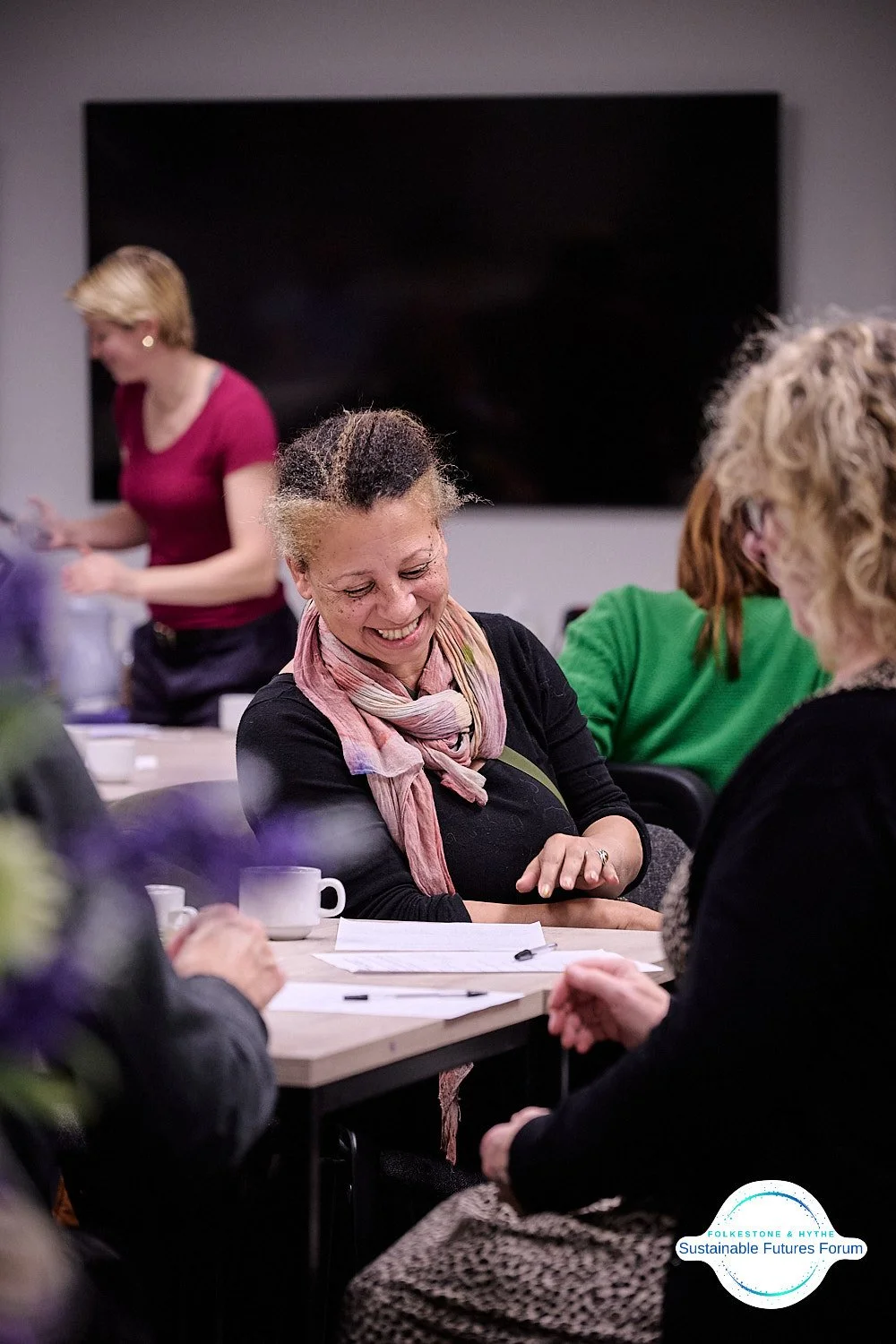 A woman smiling at a meeting or workshop, sitting at a table with papers and a coffee mug, in a room with other women and a person walking in the background.