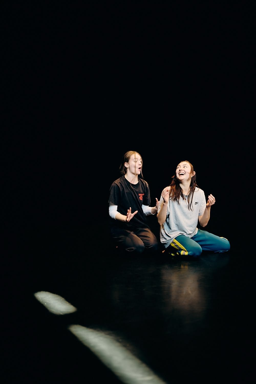 Two women sitting on a dark stage, laughing and engaging in an expressive conversation.