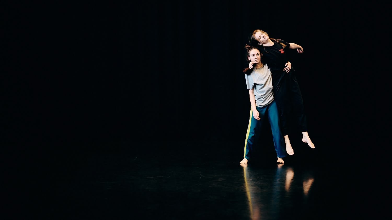 Two dancers practicing on a black stage, one lifting the other, both in casual dance attire.