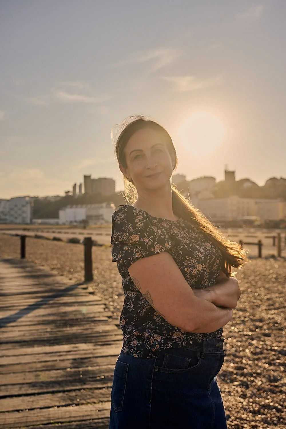 A woman standing on a wooden pathway on a beach during sunset, smiling with her arms crossed, with a cityscape in the background.