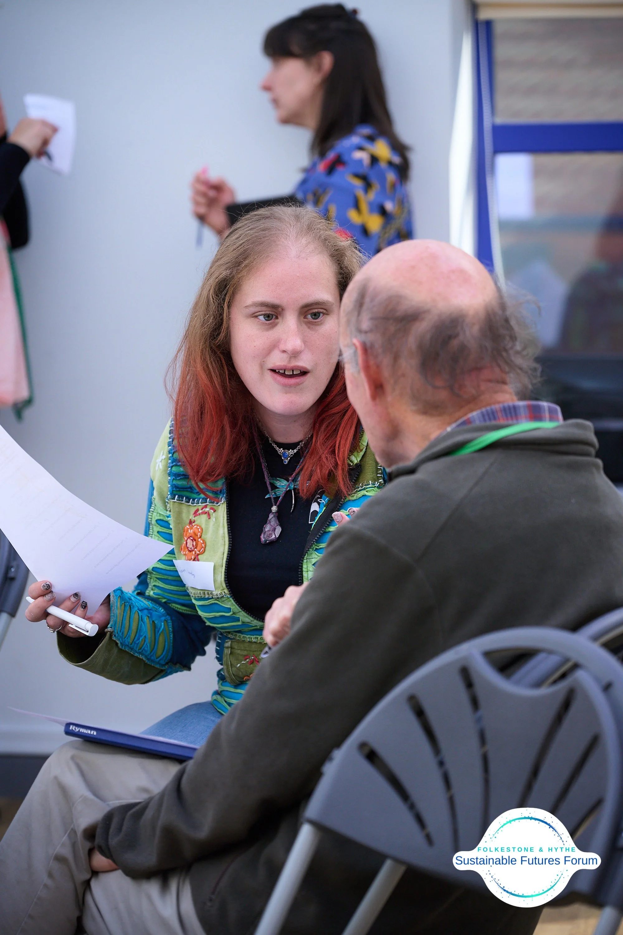 A young woman with red hair and colorful clothing is speaking with an older man with glasses at a forum, with a logo for 'Folkestone & Hythe Sustainable Futures Forum' in the corner.