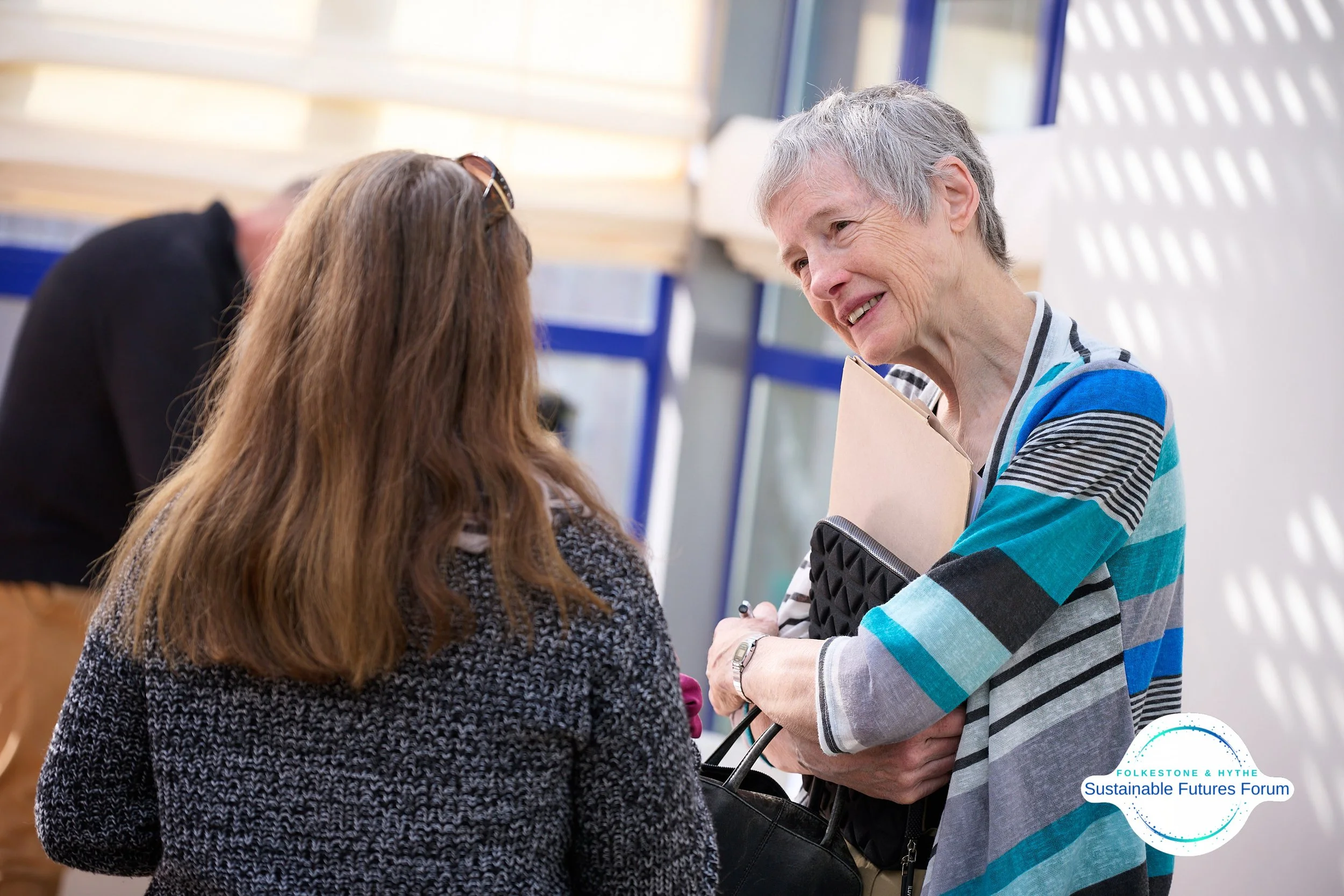 An older woman with gray hair, wearing a striped blue and gray sweater, smiling and talking to a younger woman with long brown hair, in a gray textured sweater, during a forum or conference. There is a logo for Folketone & Hythe Sustainable Futures F