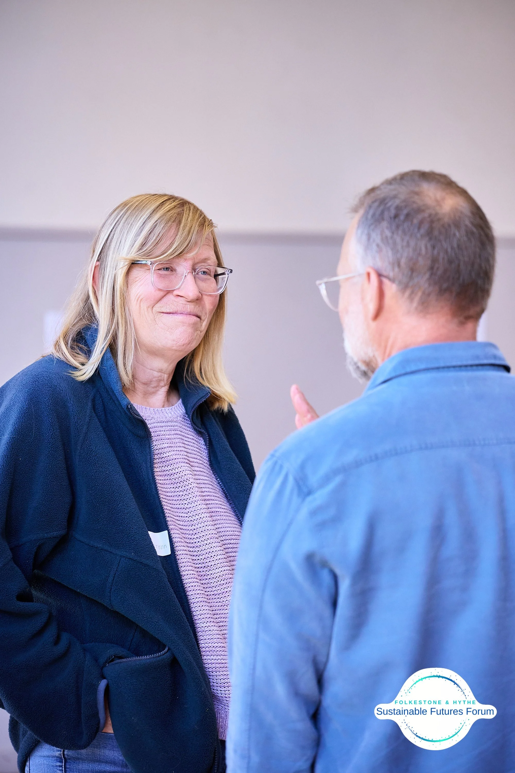 A woman and a man are engaged in conversation at the Folkestone & Hythe Sustainable Futures Forum. The woman is smiling and wearing glasses, a dark jacket, and a light purple top. The man has gray hair and a beard, is also wearing glasses and a blue 