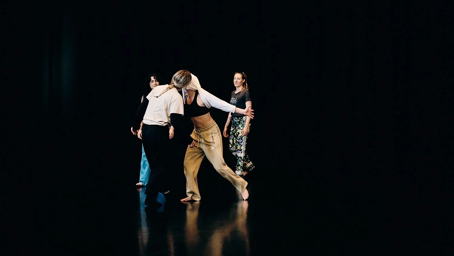 Four people dancing on a dark stage, with one person in the center holding another person, surrounded by two women observing.
