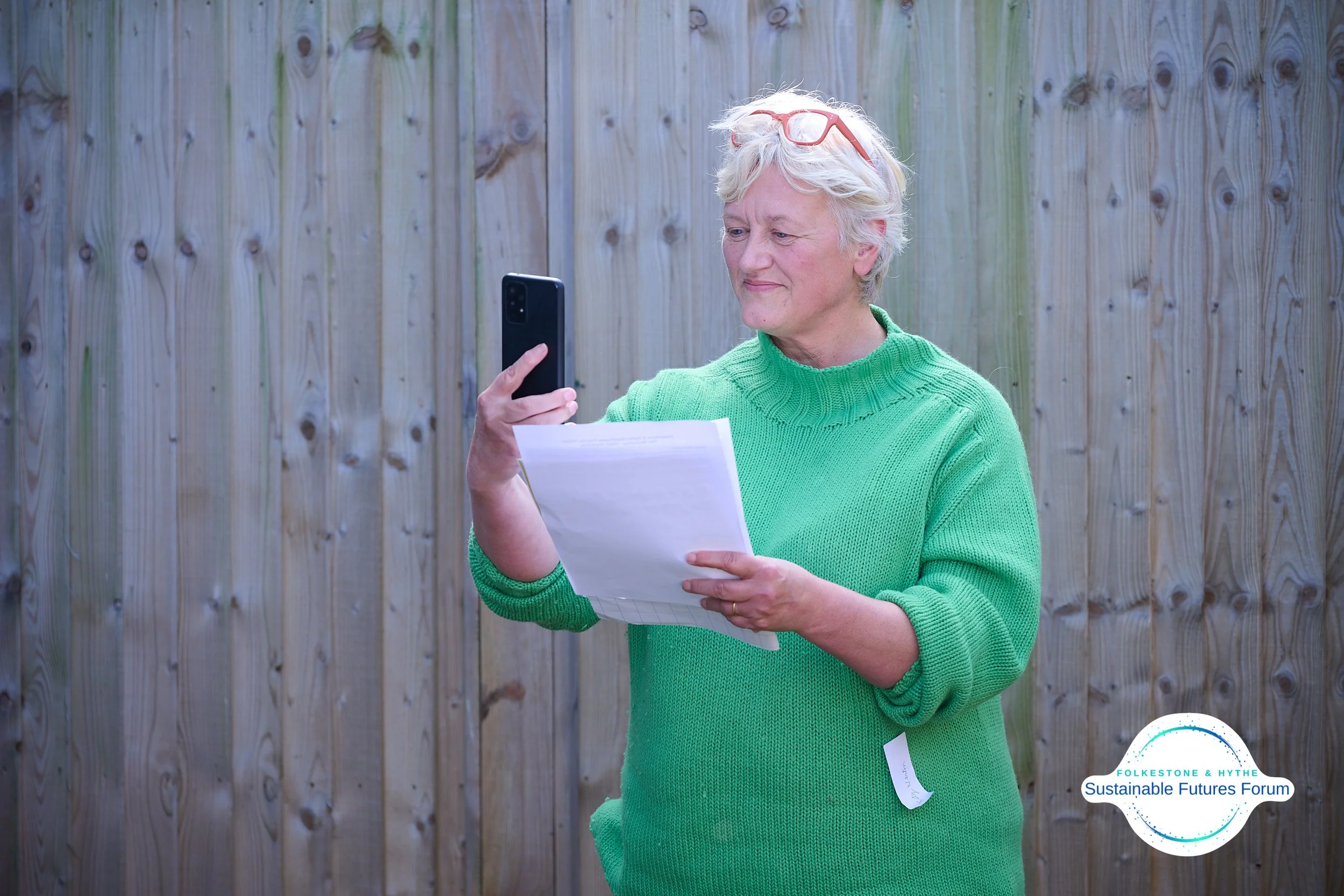 An elderly woman with short white hair, wearing a green sweater and glasses on her head, standing outdoors in front of a wooden fence, taking a photo with her smartphone while holding papers in her other hand. There is a logo in the bottom right corn