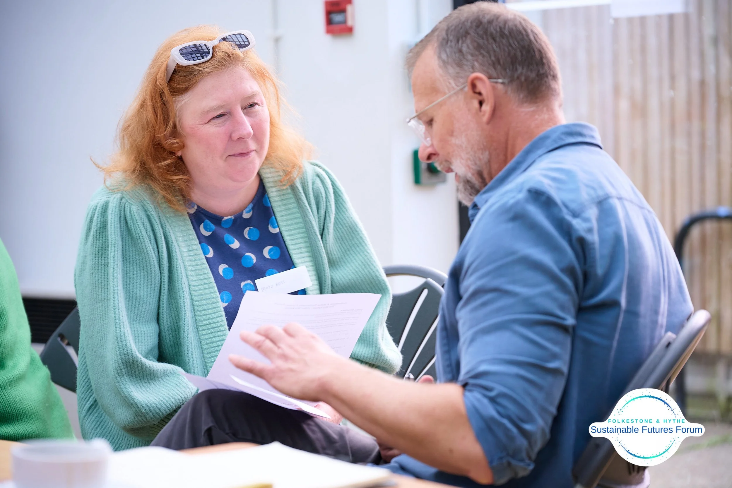 A woman with red hair, wearing sunglasses on her head and a turquoise cardigan, looks at a man with glasses and a gray beard, who is showing her some papers at a table during an outdoor event.