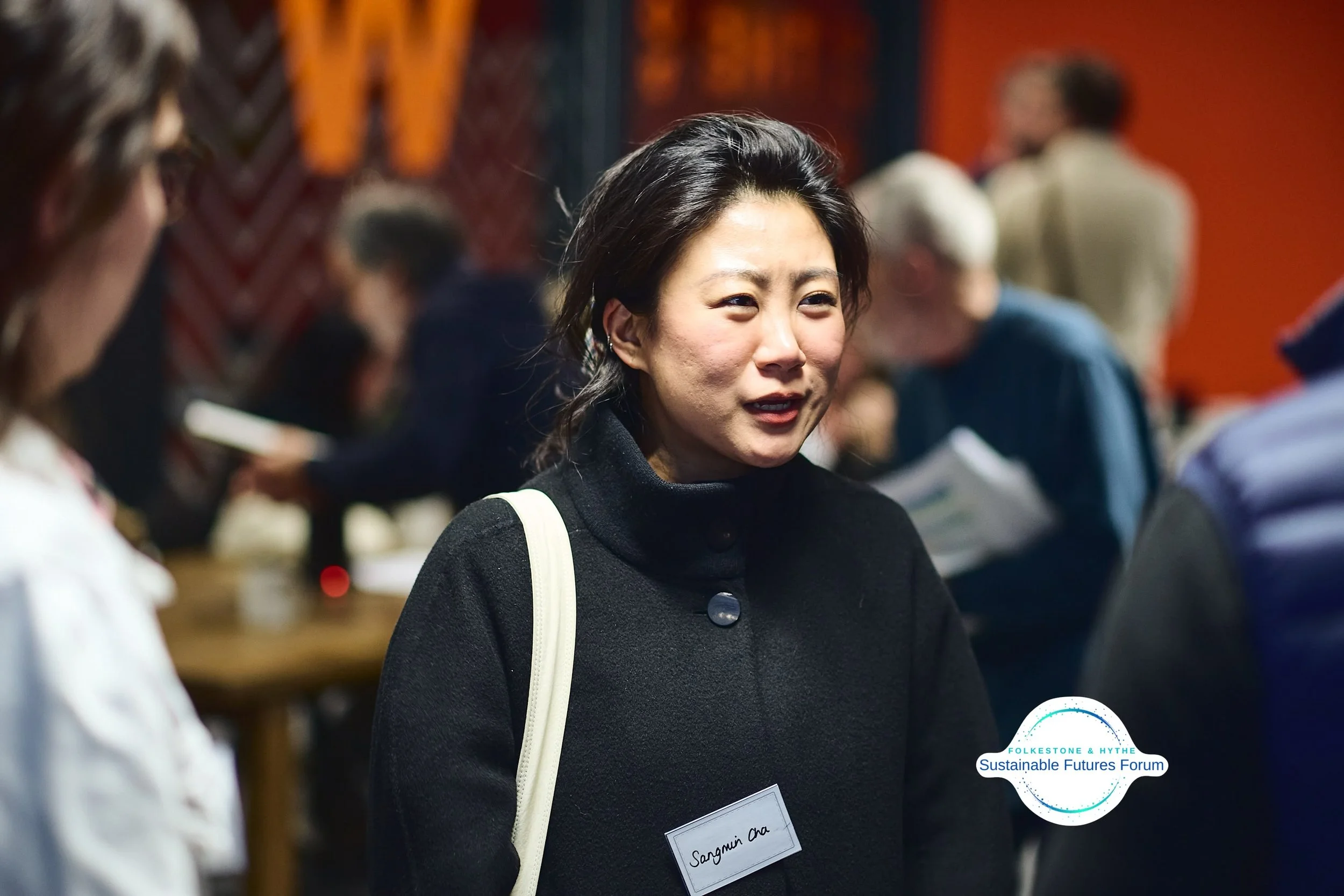 A woman with dark hair wearing a black turtleneck and a name tag at a professional event, engaging in conversation with other attendees. There is a logo for the Folkeystone & Hythe Sustainable Futures Forum in the corner.