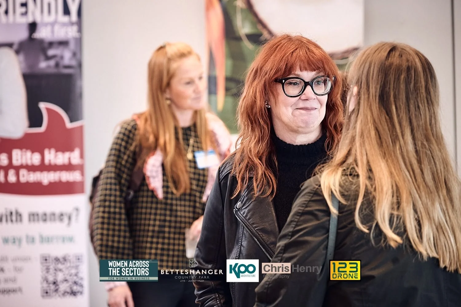Two women with red hair and glasses engaged in conversation at an indoor event, with a third woman in the background near a poster.