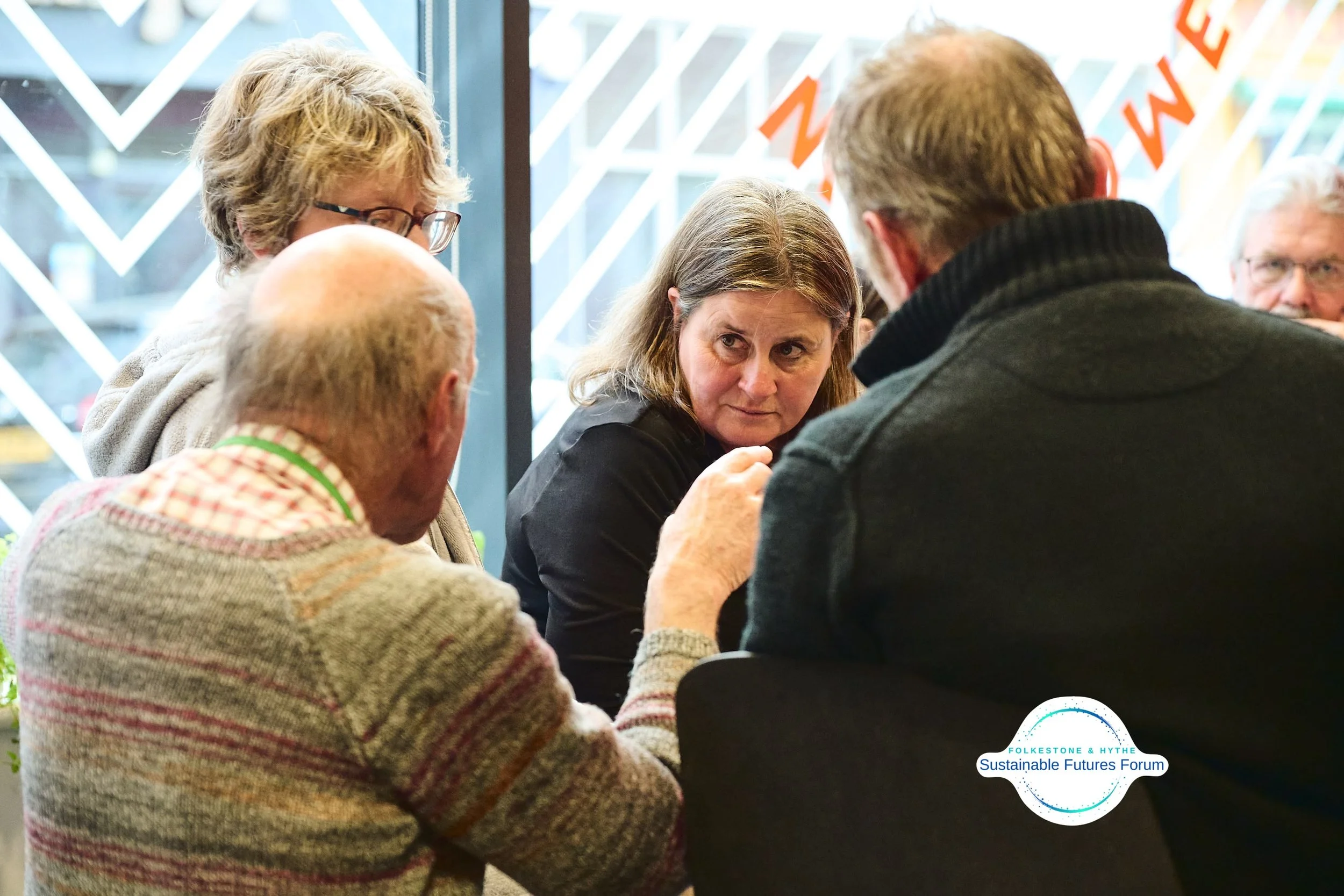 A group of five older adults sitting around a table engaged in a discussion at an indoor event, with a window and patterned decorations in the background, and a logo for Folkestone & Hythe Sustainable Futures Forum.
